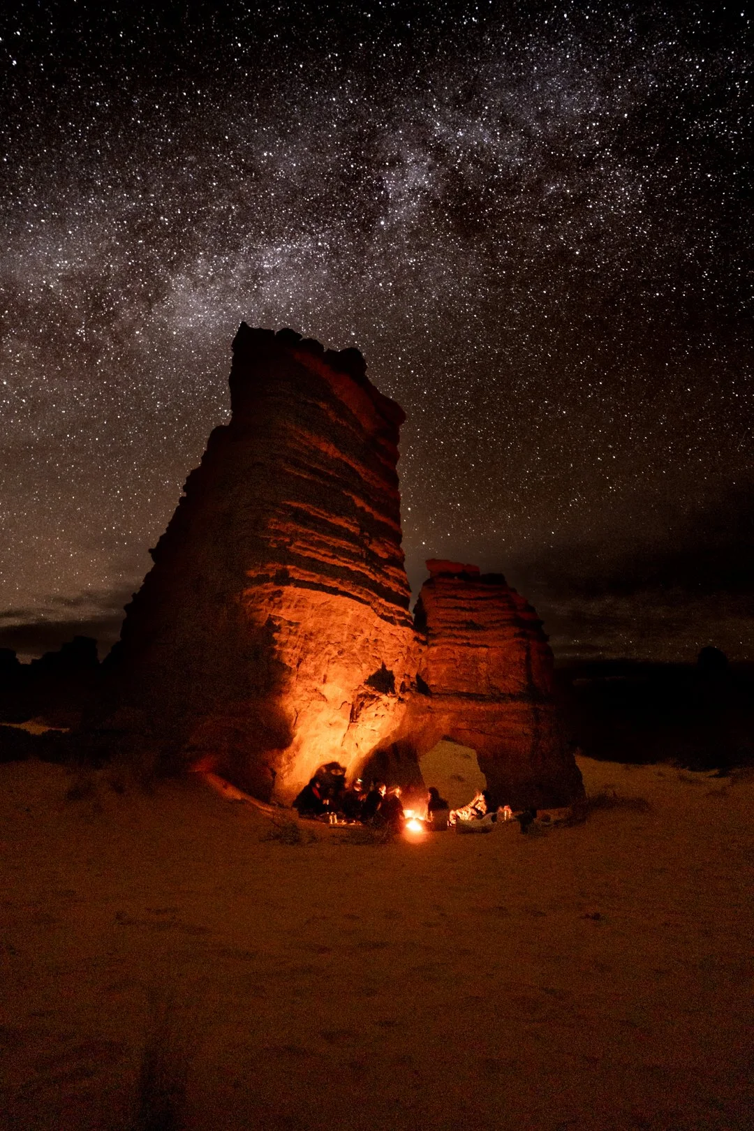 A nighttime desert scene with two large rock formations illuminated by warm light and a group of people sitting by a campfire at the base, under a star-filled sky on Tassili n'Ajjer Plateau in the Sahara desert, Algeria.
