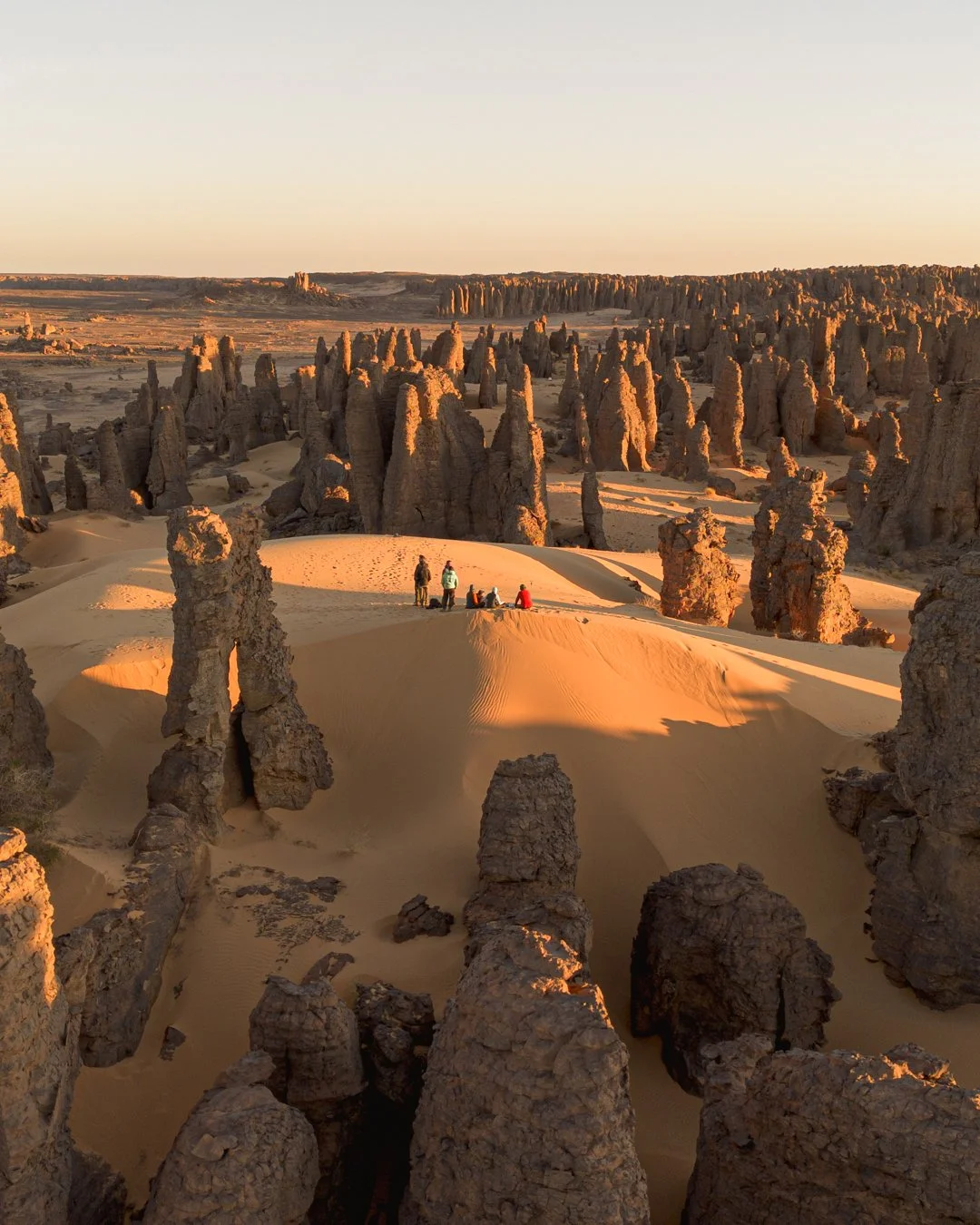 A desert landscape with numerous tall, spire-shaped rock formations and a few people sitting on a sandy hill at sunrise on Tassili n'Ajjer Plateau in the Sahara desert, Algeria