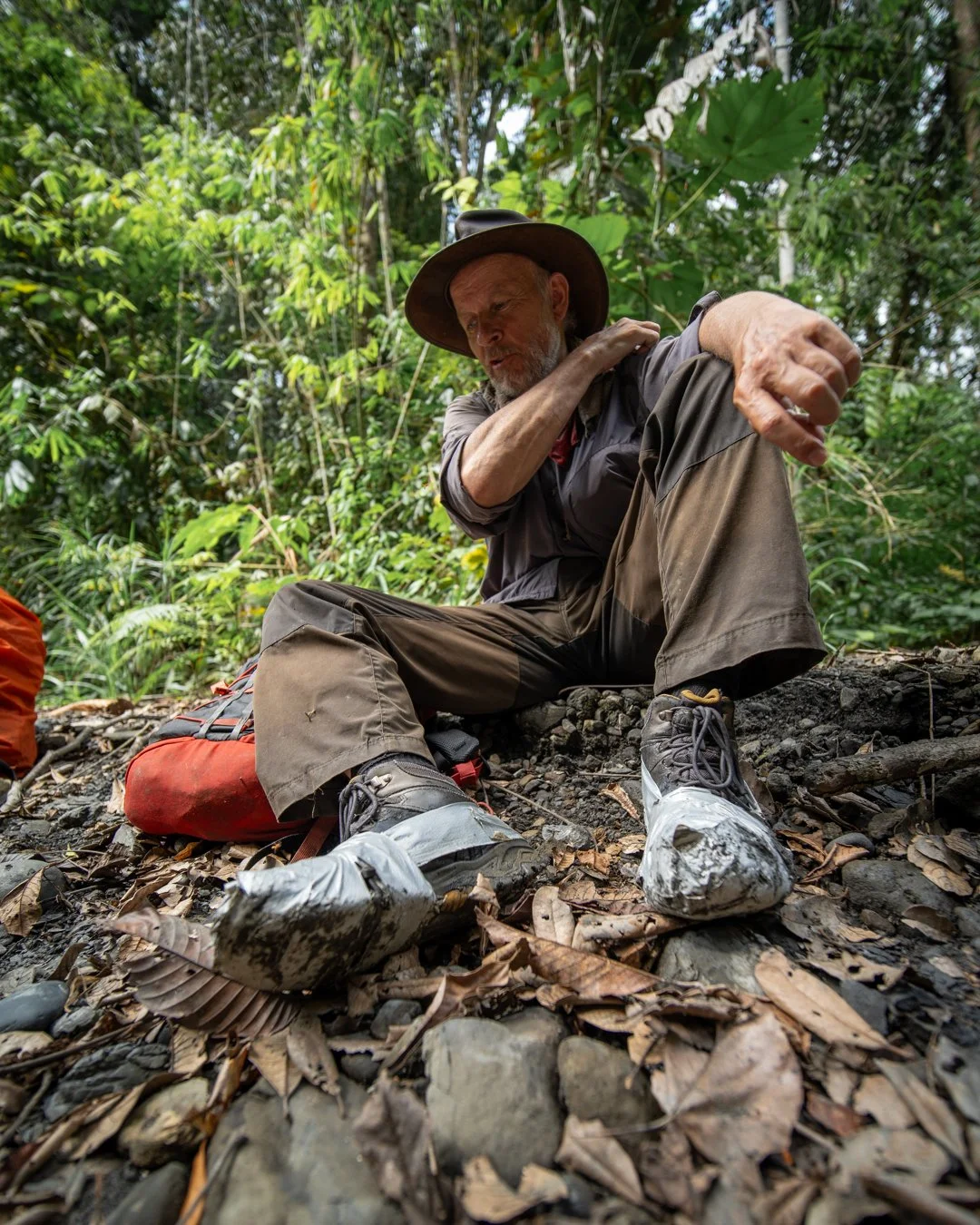 A man sitting on a forest floor with leaves and rocks, wearing a wide-brimmed hat, outdoor clothes, and broken sneakers repaired with duct tape, with a red backpack nearby, surrounded by dense green jungle vegetation.