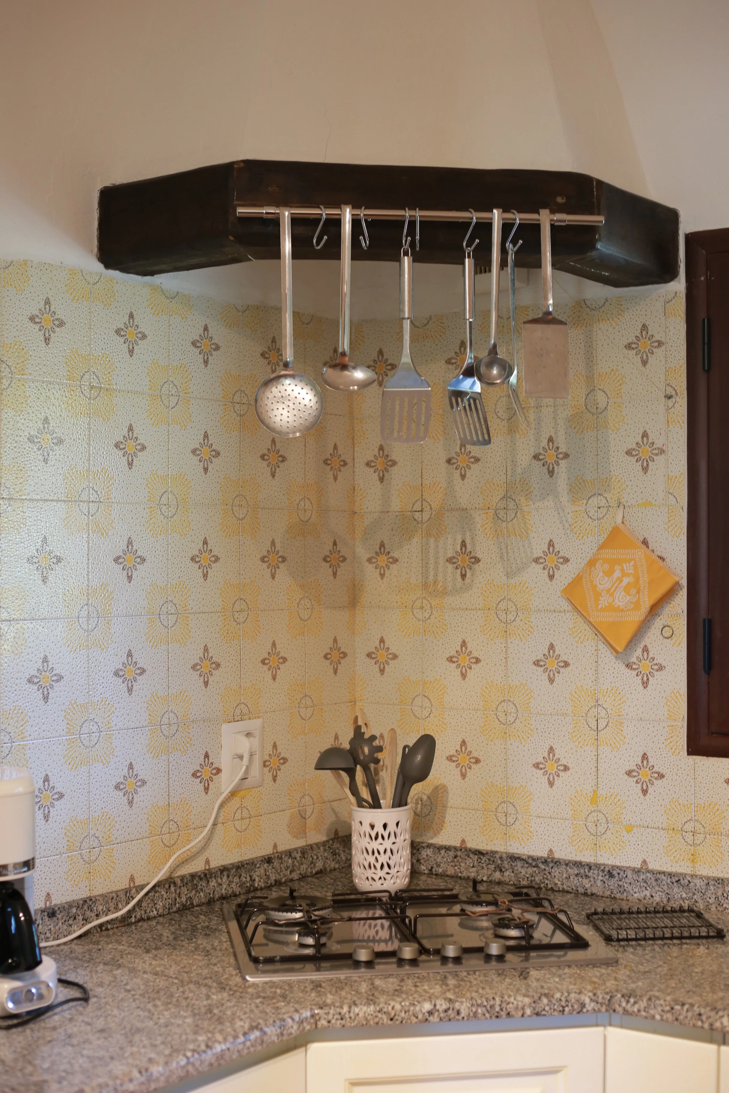 Küche mit bunter, blumiger Fliesenspiegel, Gasherd, einen weißen Utensilienkorb mit Kochutensilien auf der Arbeitsplatte, elektrische Steckdose an der Wand und eine Holz-Wandverkleidung