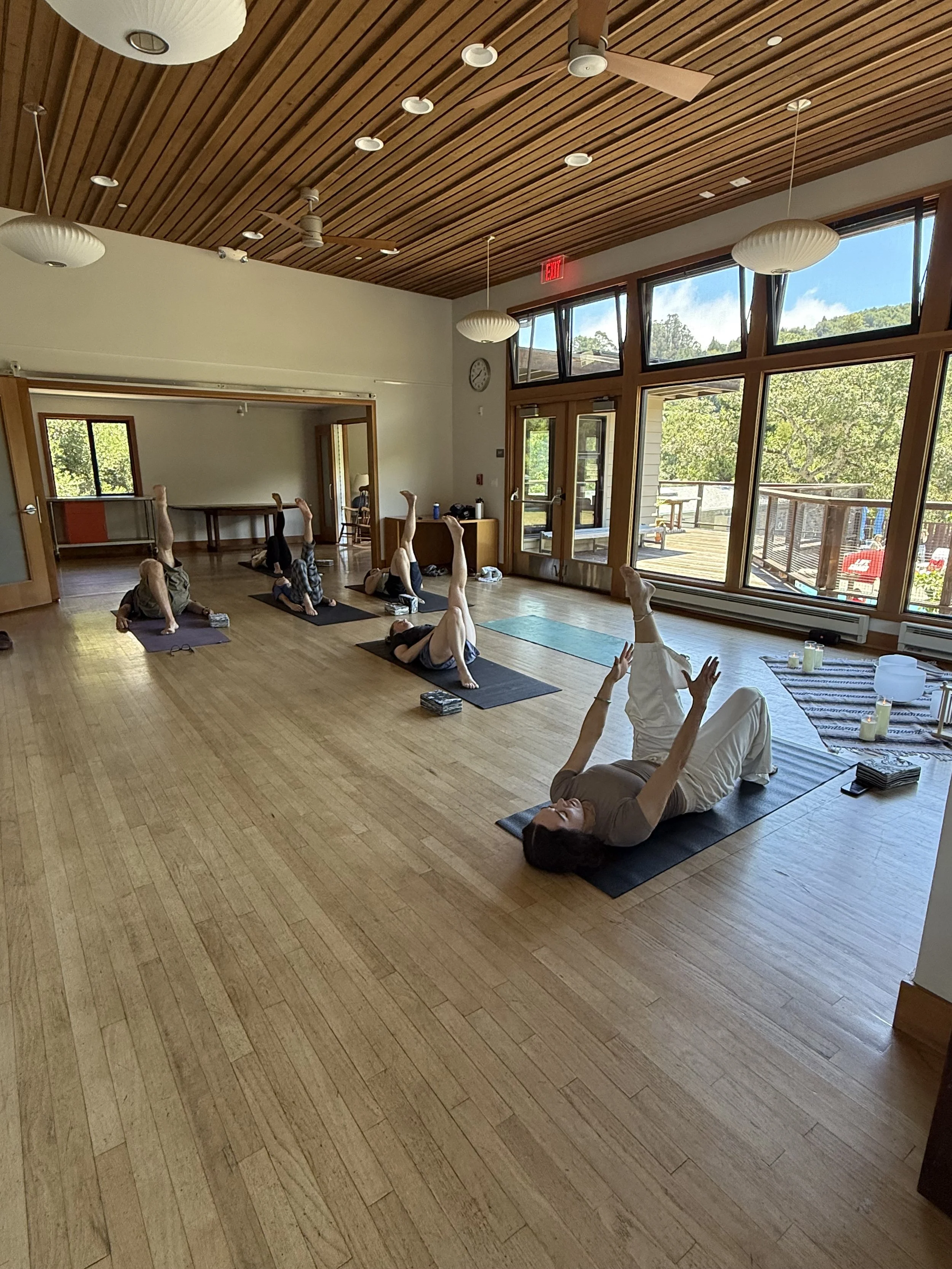 People participating in a yoga class in a bright room with large windows, wooden ceiling, and hardwood floors. They are lying on yoga mats with one leg raised, practicing a pose.