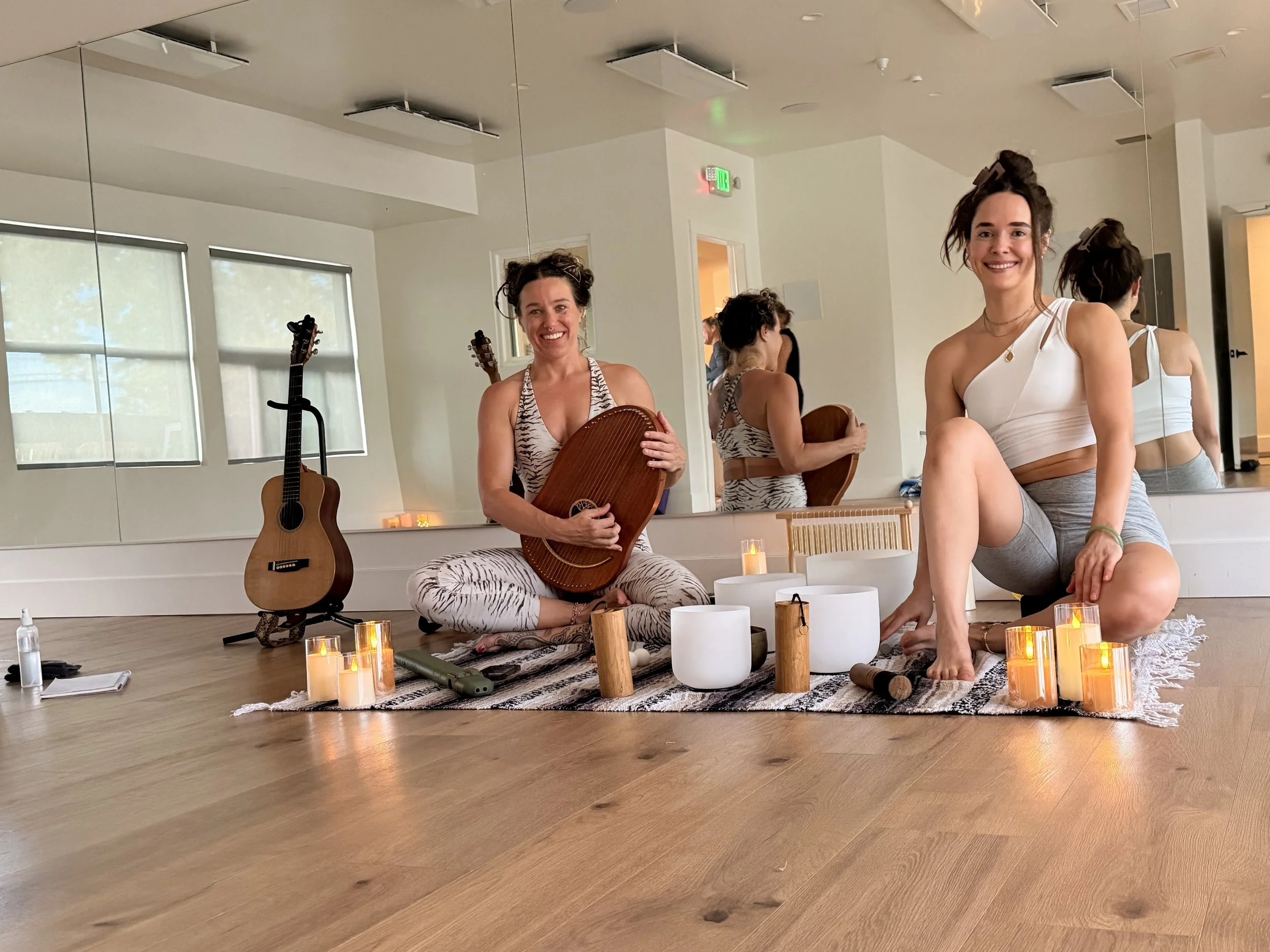 Two women with long dark hair, yoga balls, and yoga mats are in a modern room with white walls and natural light. One woman is smiling while sitting on the floor with her. The other woman is smiling while sitting on a yoga ball. There are candles, a 