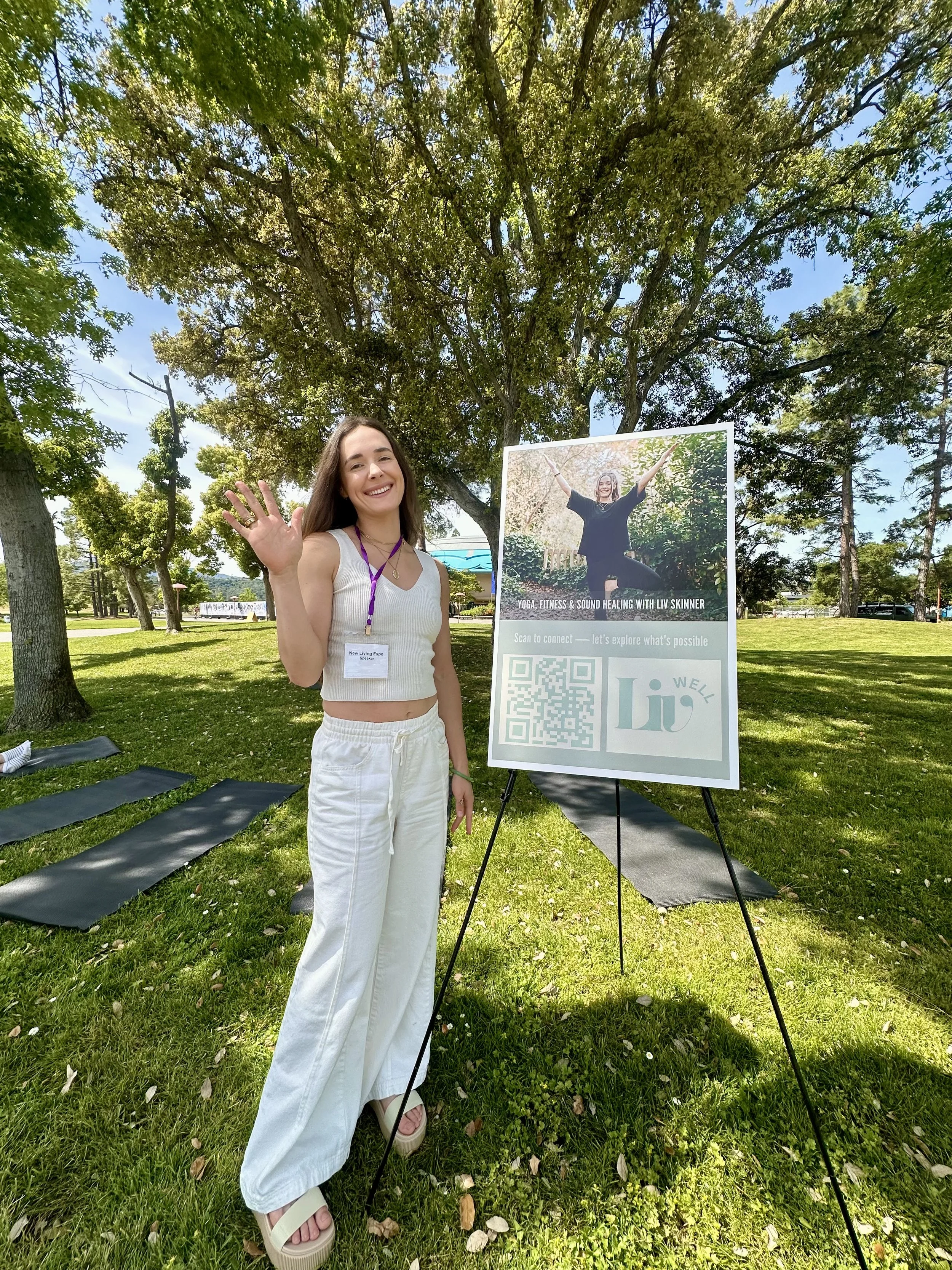 A smiling woman wearing a white tank top and white pants, waving at the camera, standing on a grassy park with trees and a stand-up poster that promotes yoga, fitness, and sound healing with Liv Skinner.