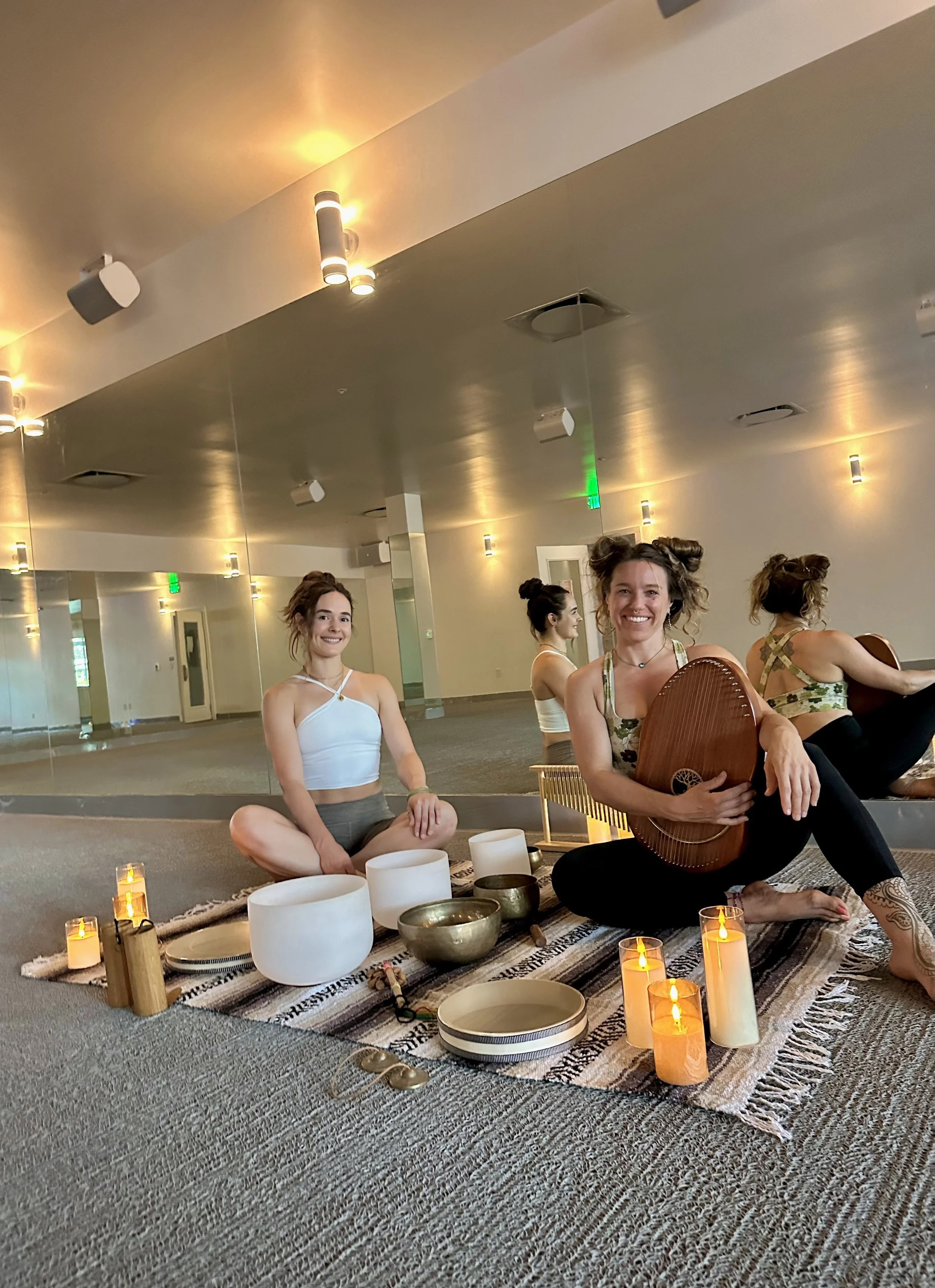 Group of women practicing yoga or meditation in a room decorated with candles and sound bowls, sitting on a patterned rug.