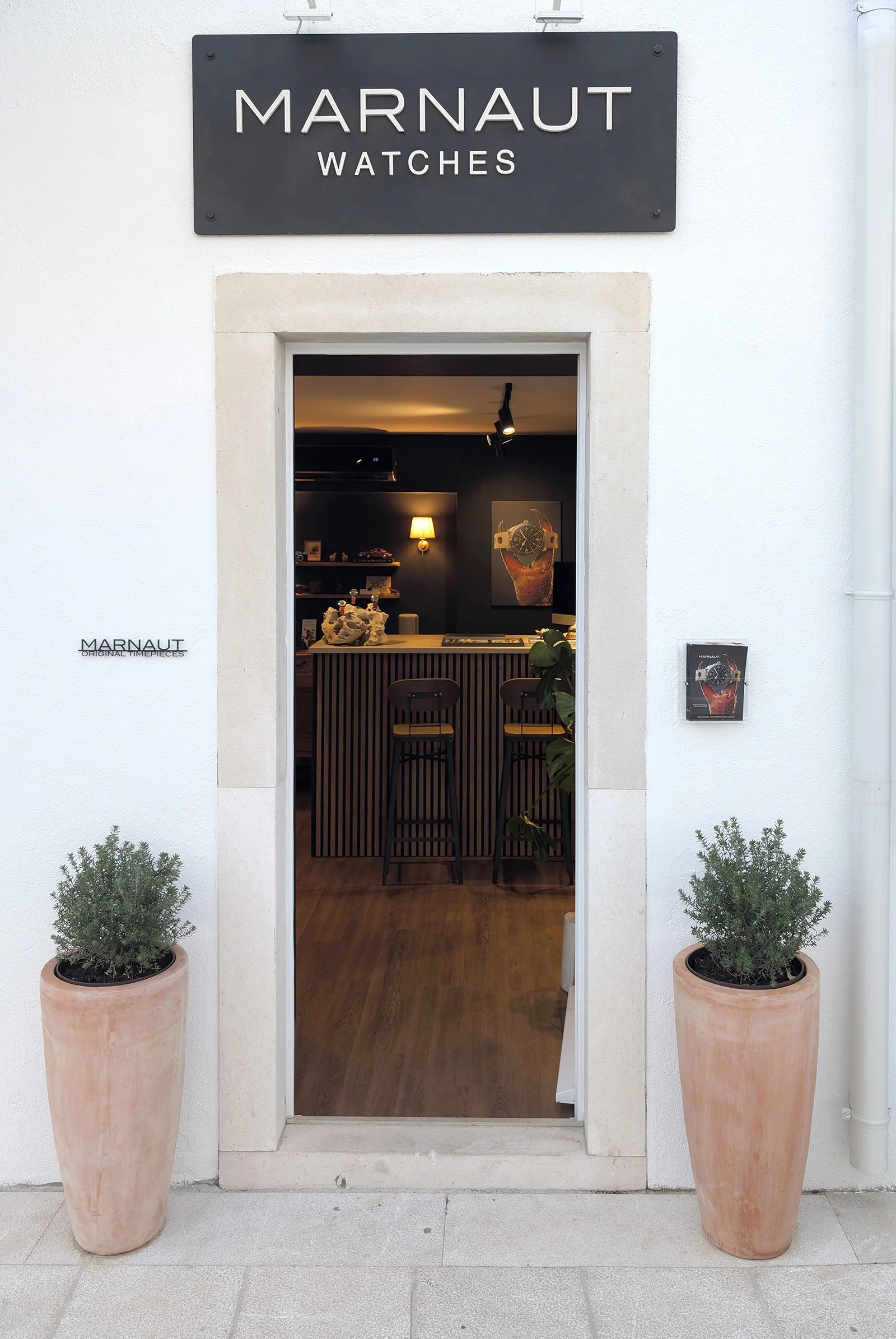 Storefront with a black sign reading 'Marnaut Watches' above a doorway. Two large terracotta pots with green plants flank the entrance, and inside, there is a dark-colored interior with wooden furniture, a wall-mounted lamp, and a wristwatch advertisement on the wall.
