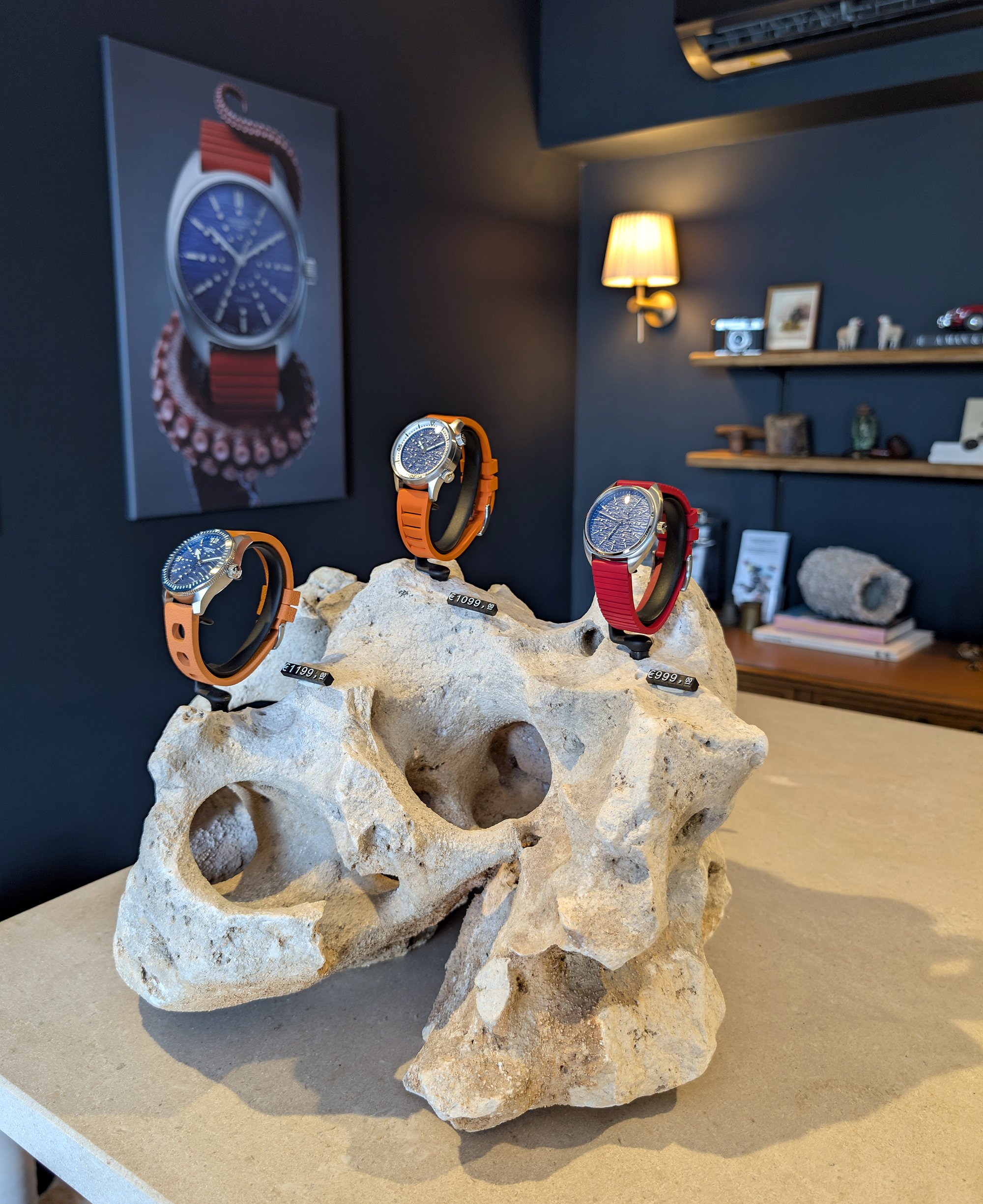 Multi-colored wristwatches displayed on a rock formation in a store with dark blue walls, featuring framed pictures, a shelf with decorative items, and a wall-mounted lamp.