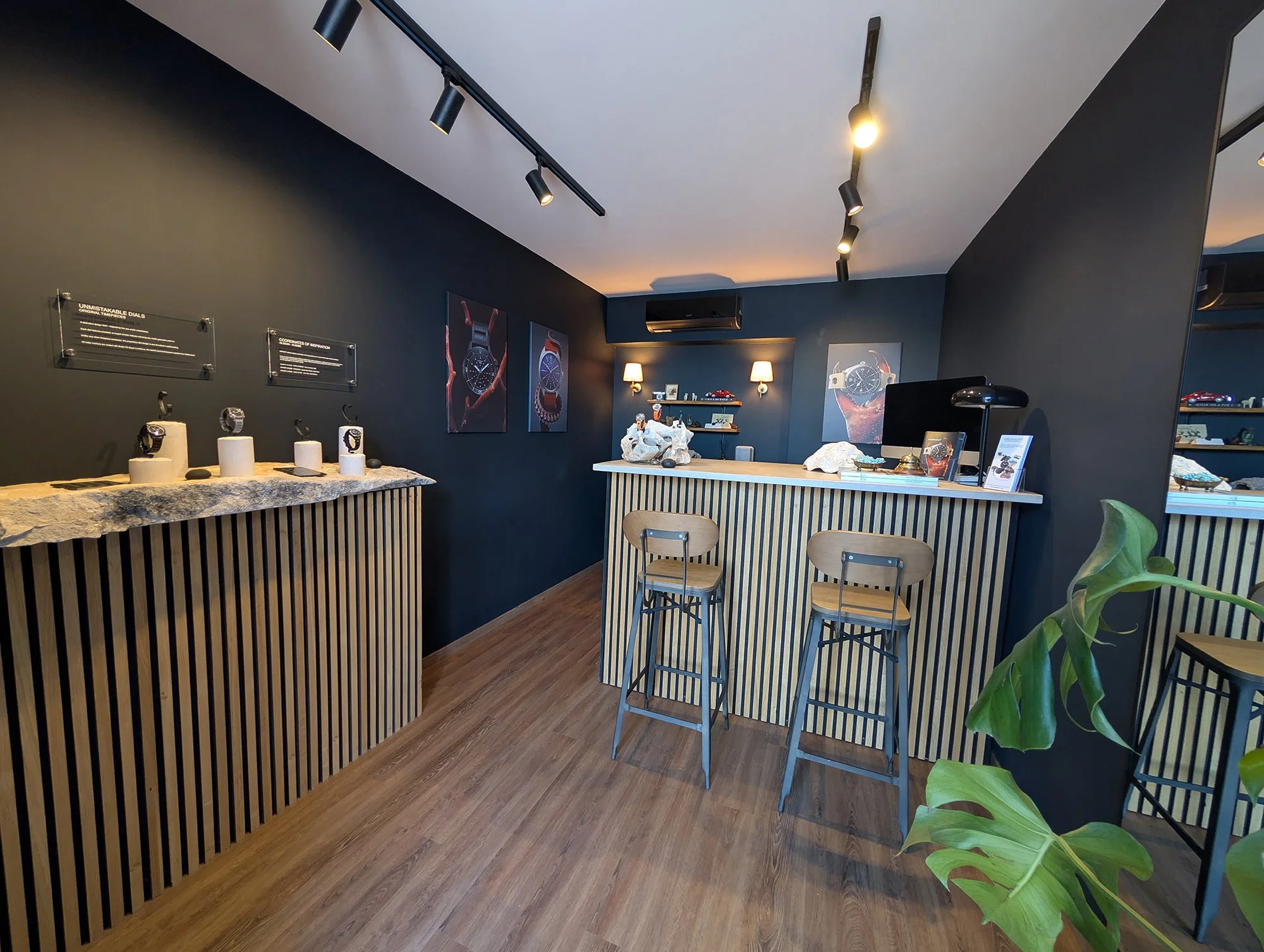 Interior of a watch showroom with dark walls and wooden flooring, featuring display tables with watches, framed watch posters, a counter with books and decorative rocks, wall-mounted lamps, and a large mirror on the right side.