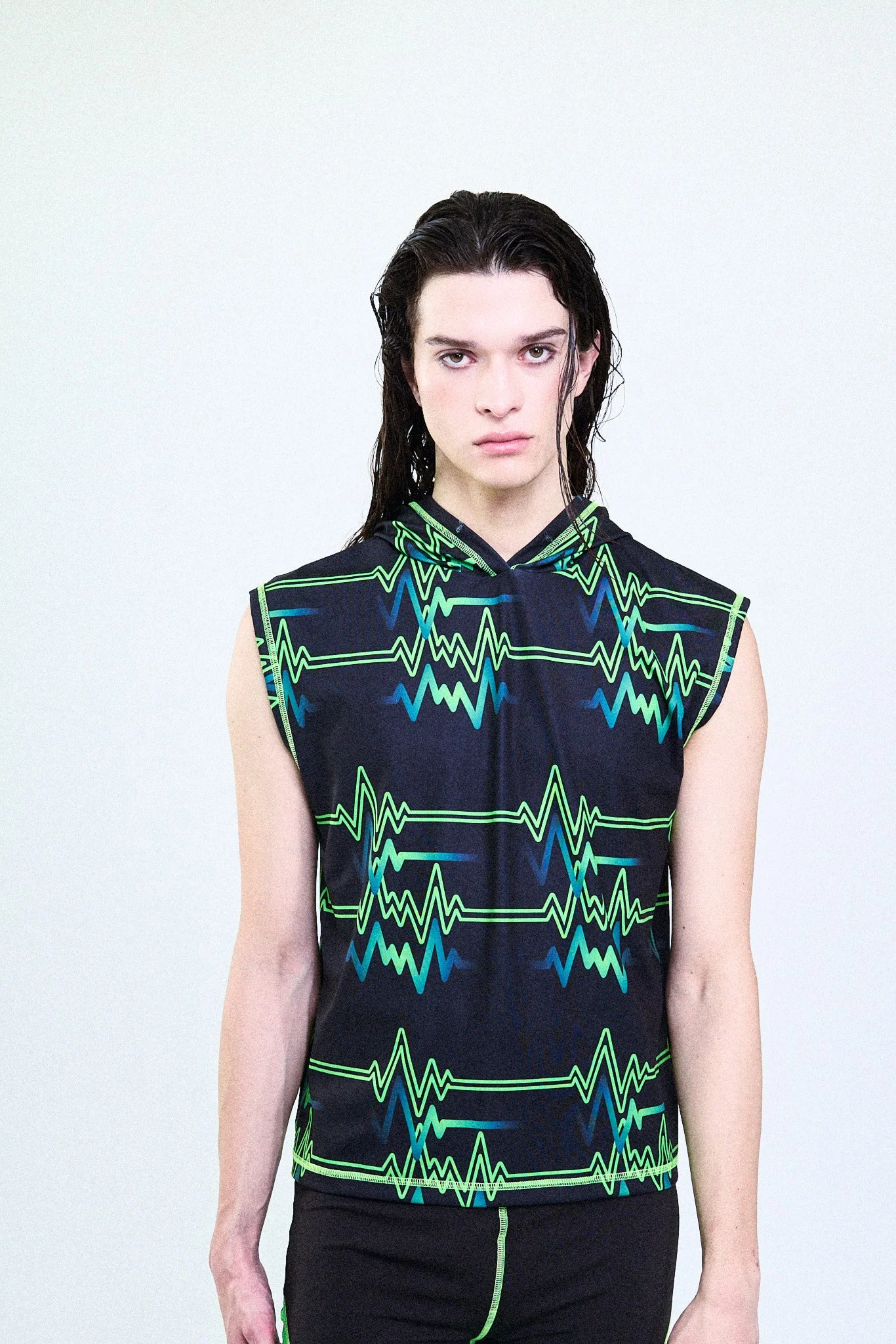 A person with long black hair and light skin wearing a sleeveless black athletic shirt with green, blue, and teal heartbeat line patterns, standing against a plain white background.