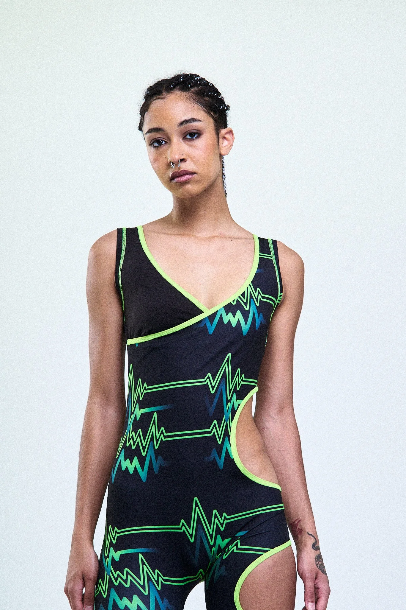 A young woman with braided hair and multiple facial piercings wearing a black athletic bodysuit with neon green and blue electric circuit line patterns, standing against a plain white background.
