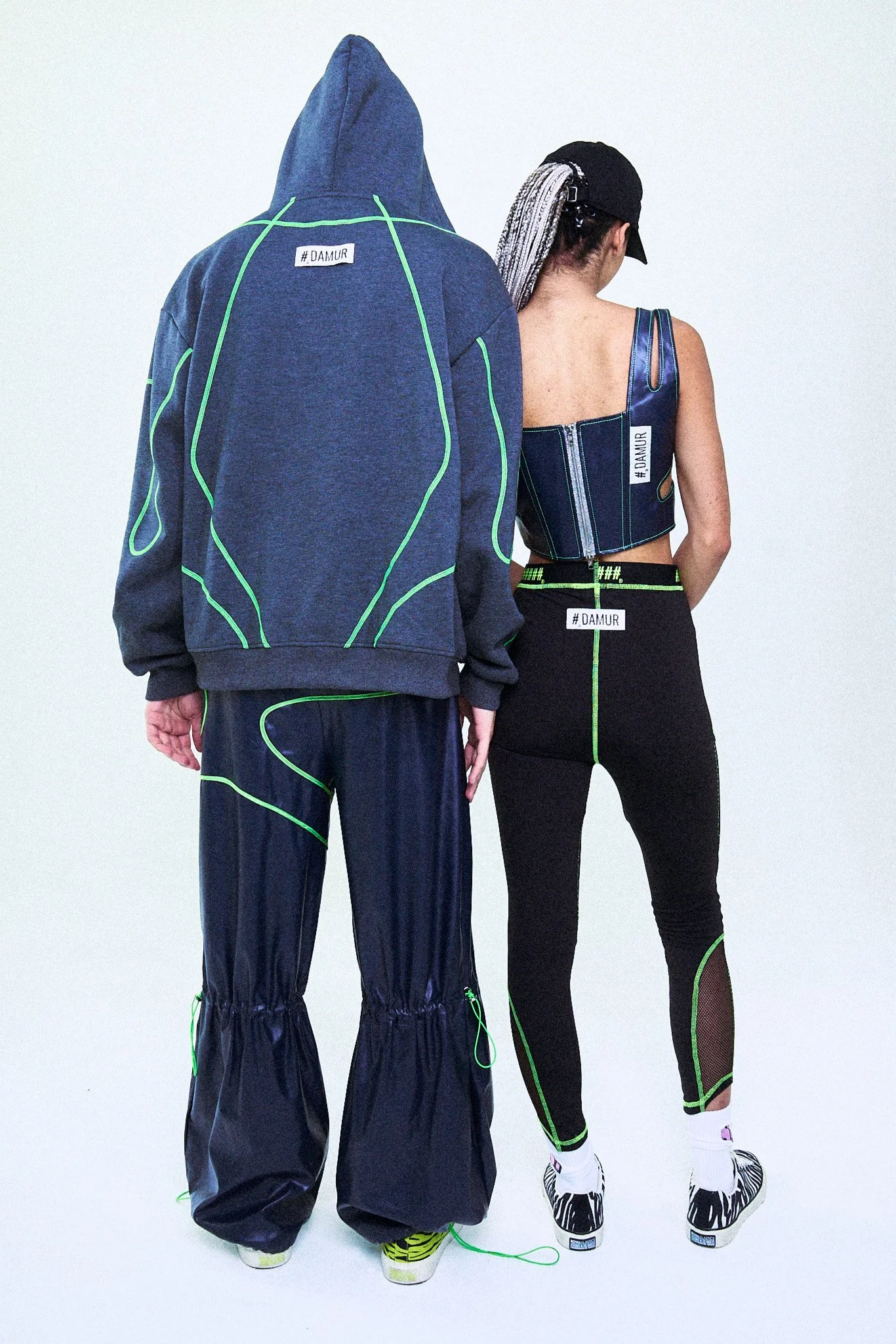 Man and woman dressed in black athletic wear with green accents, standing with their backs to the camera, against a plain white background.