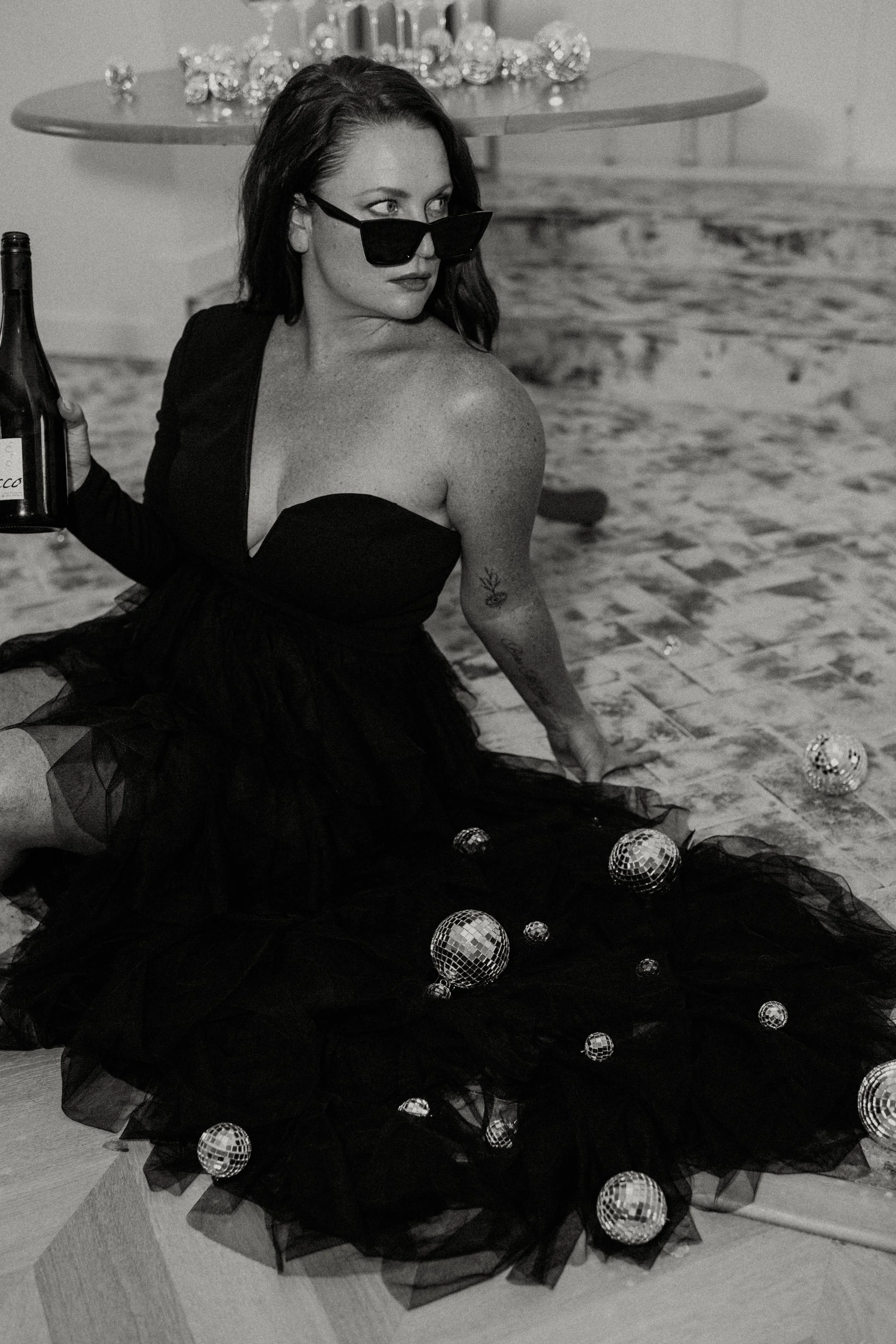 A woman with dark hair wearing black sunglasses and a strapless black tulle dress, sitting on the floor surrounded by small disco balls, holding a bottle of wine with a decorative table behind her.