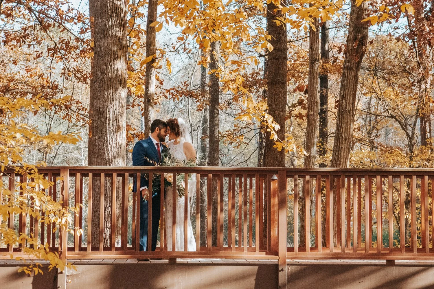 A bride and groom standing on a wooden deck surrounded by tall trees with yellow autumn leaves, sharing a moment together outdoors.