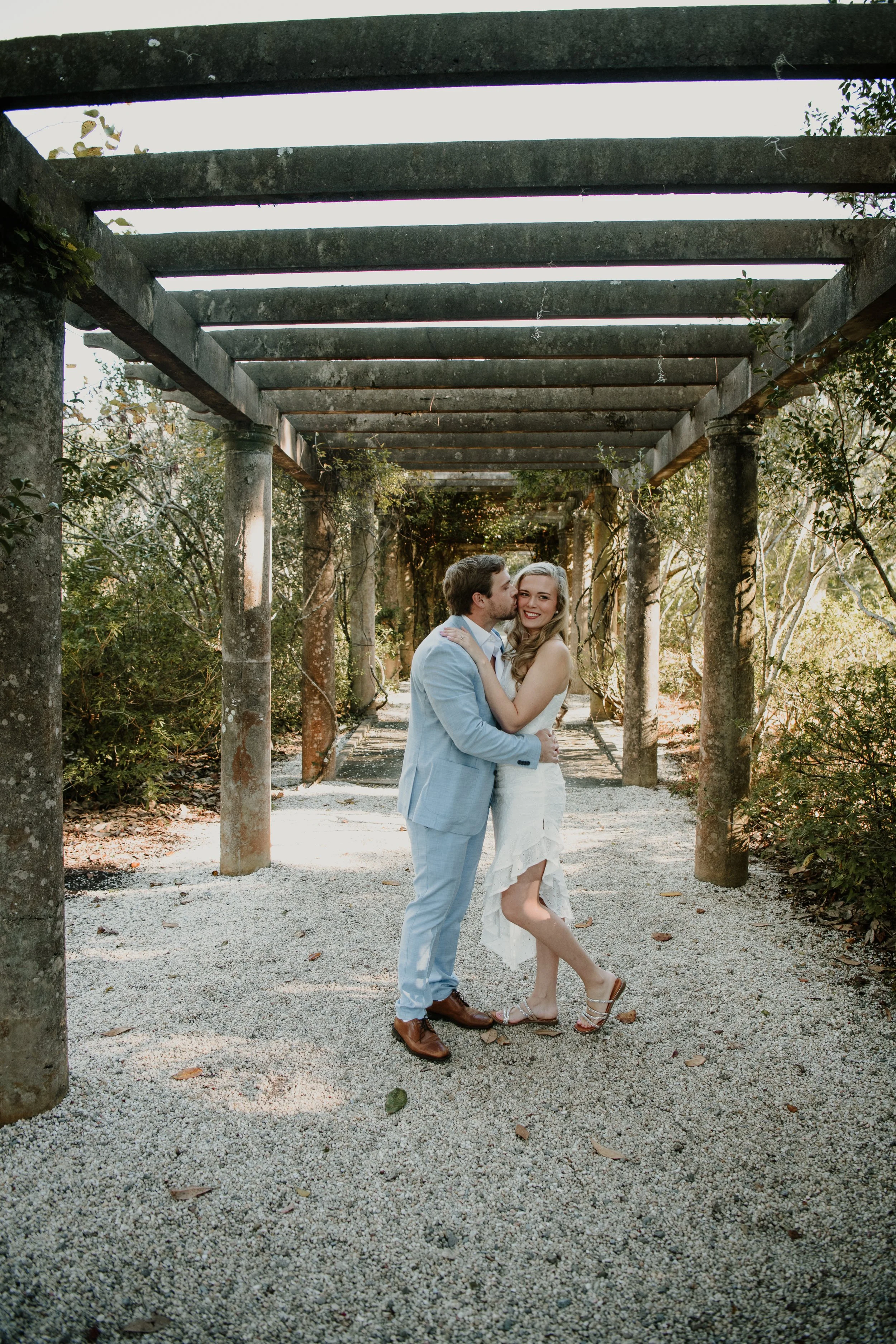 A couple in wedding attire embracing and kissing under a pergola in an outdoor setting, surrounded by greenery.