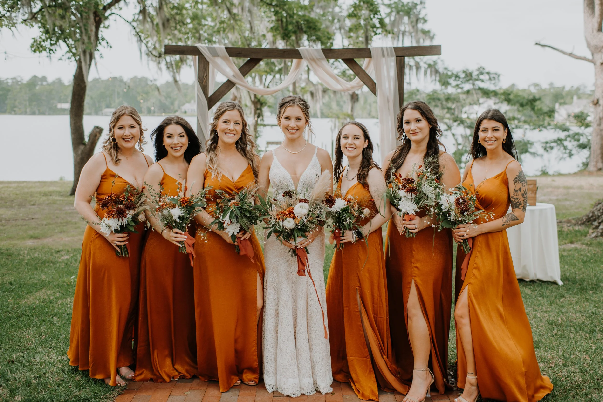Bridal party standing outdoors during a wedding, with seven women dressed in satin rust-colored dresses and holding bouquets of flowers, including a bride in white lace gown at center, under a wooden arch decorated with fabric, trees, and water in th