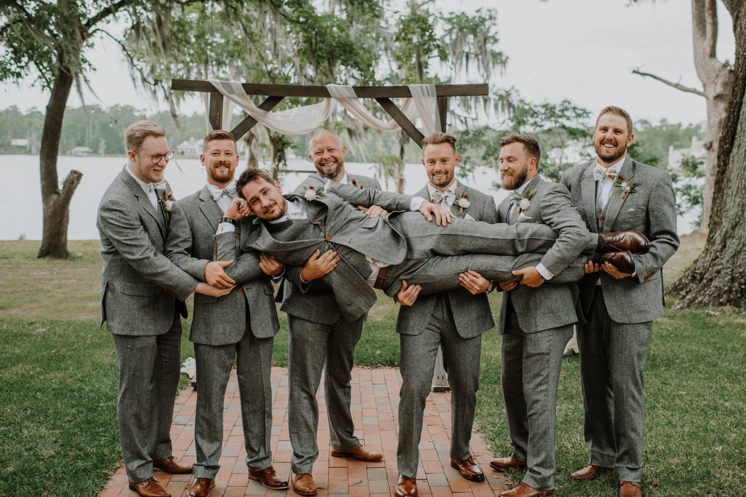 Group of seven men in gray suits holding a man horizontally at an outdoor wedding ceremony near a lake, with trees and a wooden arch decorated with white fabric in the background.