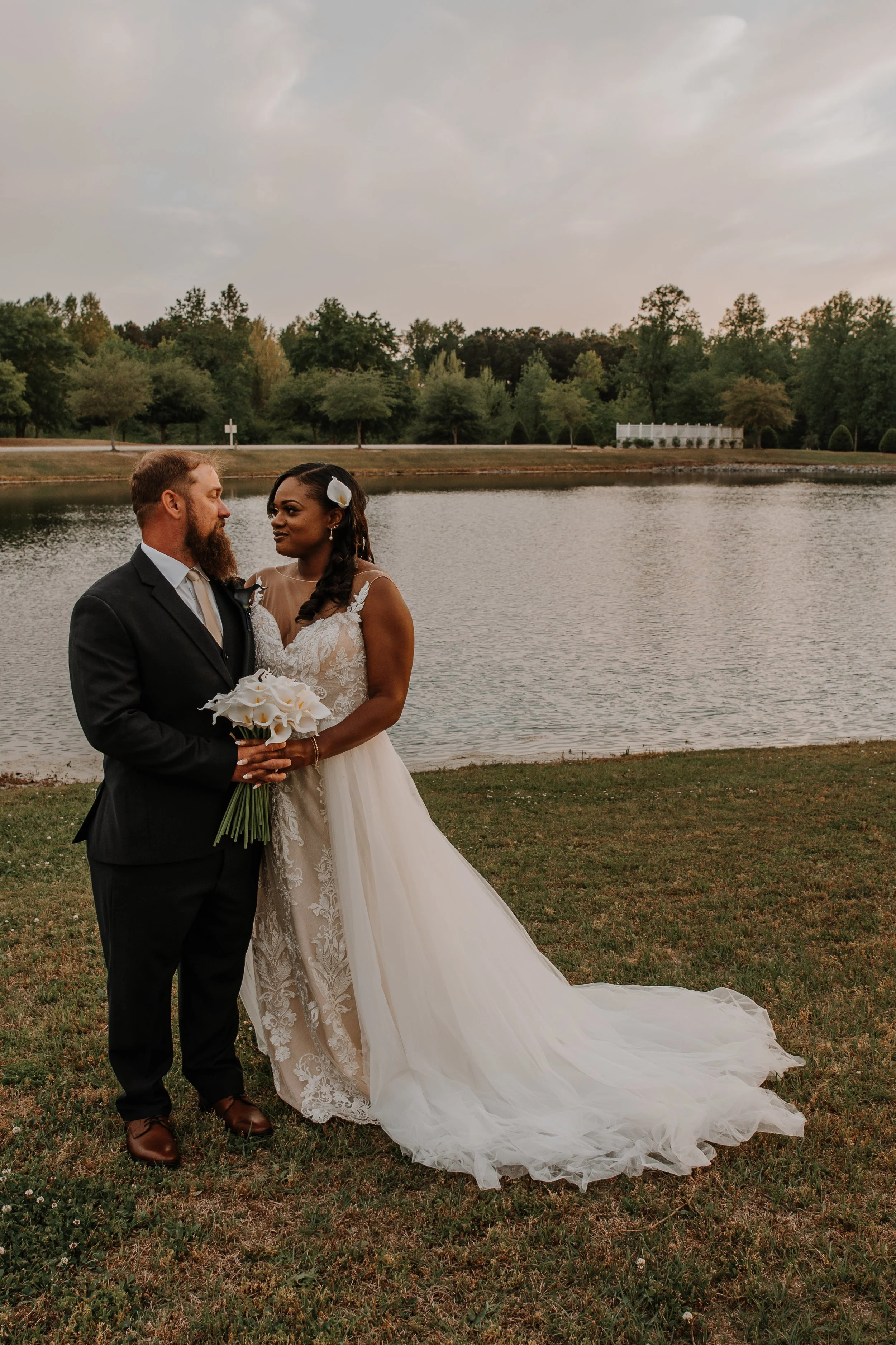 Bride and groom standing by a lake during sunset at their wedding, holding a bouquet of white calla lilies.
