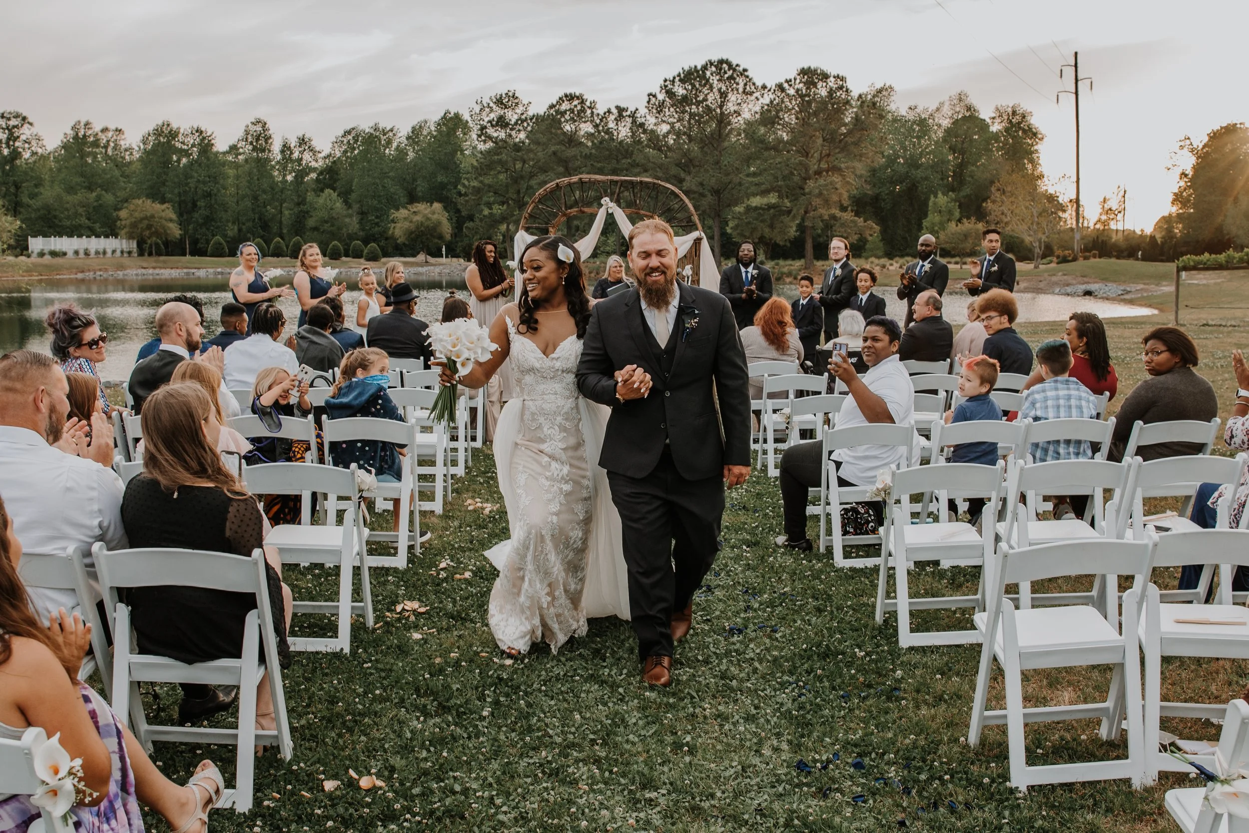 Bride and groom walking down the aisle at an outdoor wedding ceremony by a lake, surrounded by seated guests, with a decorative arch and trees in the background.