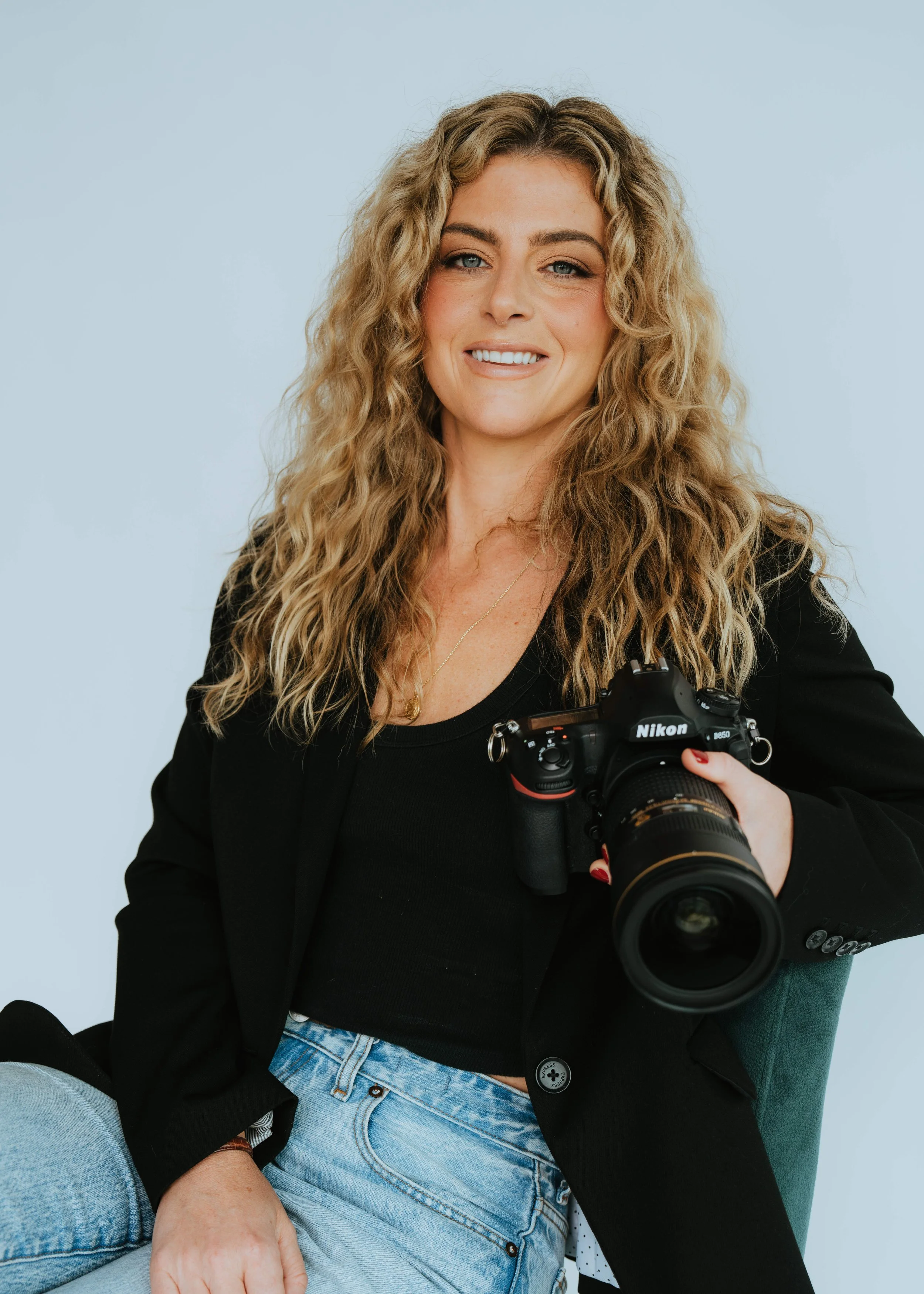 A woman with long, curly blonde hair holding a Nikon camera, smiling at the camera, wearing a black blazer and light blue jeans, sitting against a light blue background.