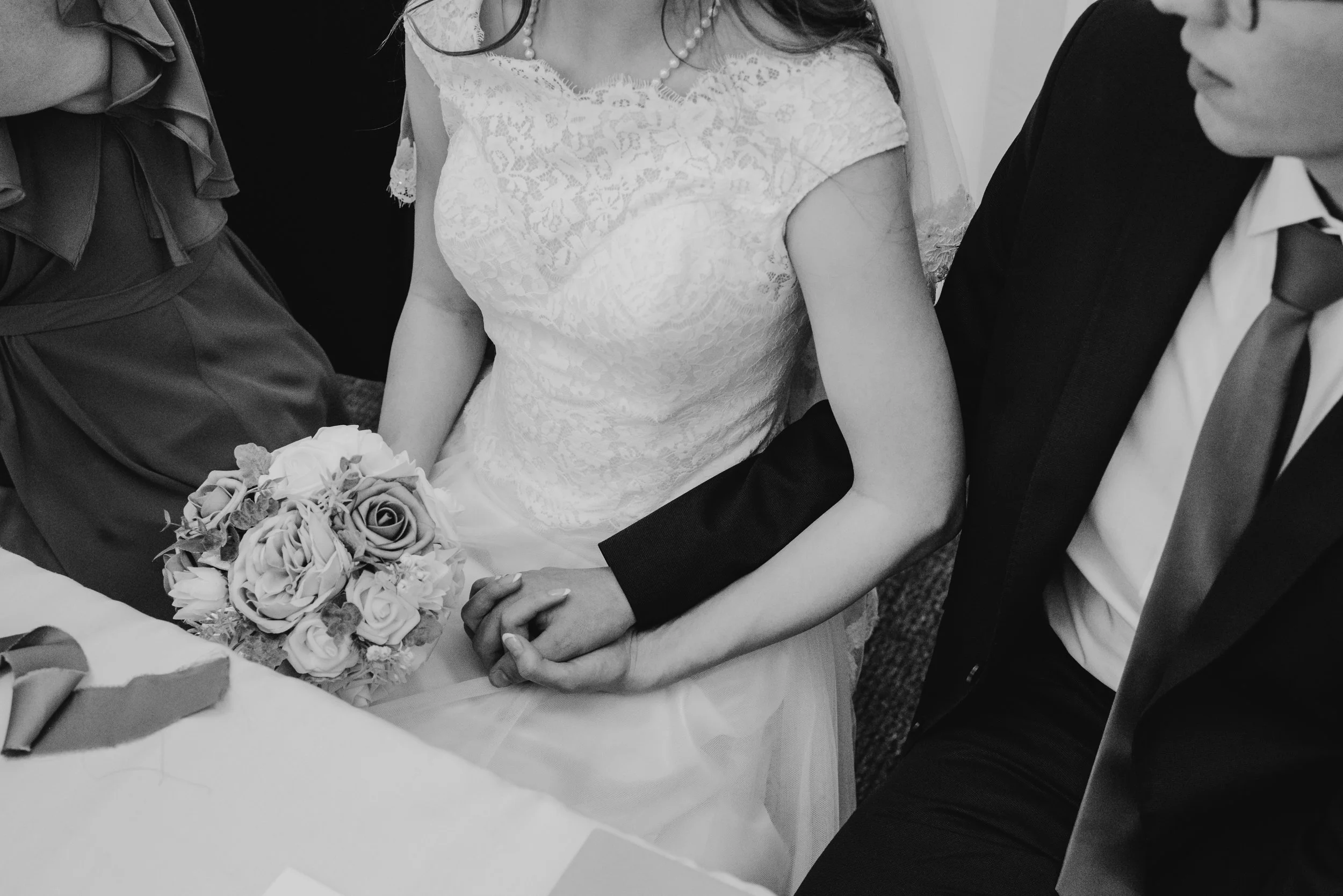 Black and white photo of a woman in a lace wedding dress holding a bouquet of flowers, sitting next to a man in a suit with a tie. Their hands are intertwined on their laps.