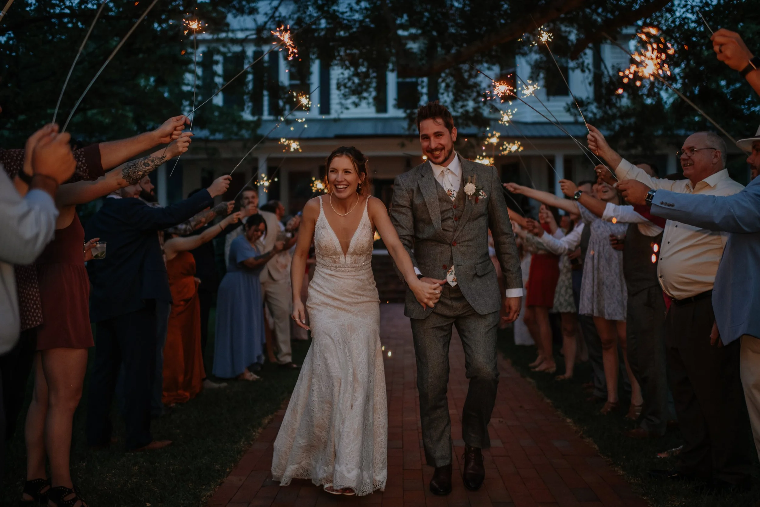 A bride and groom holding hands and smiling as they walk through a sparkler send-off crowd at their wedding reception.