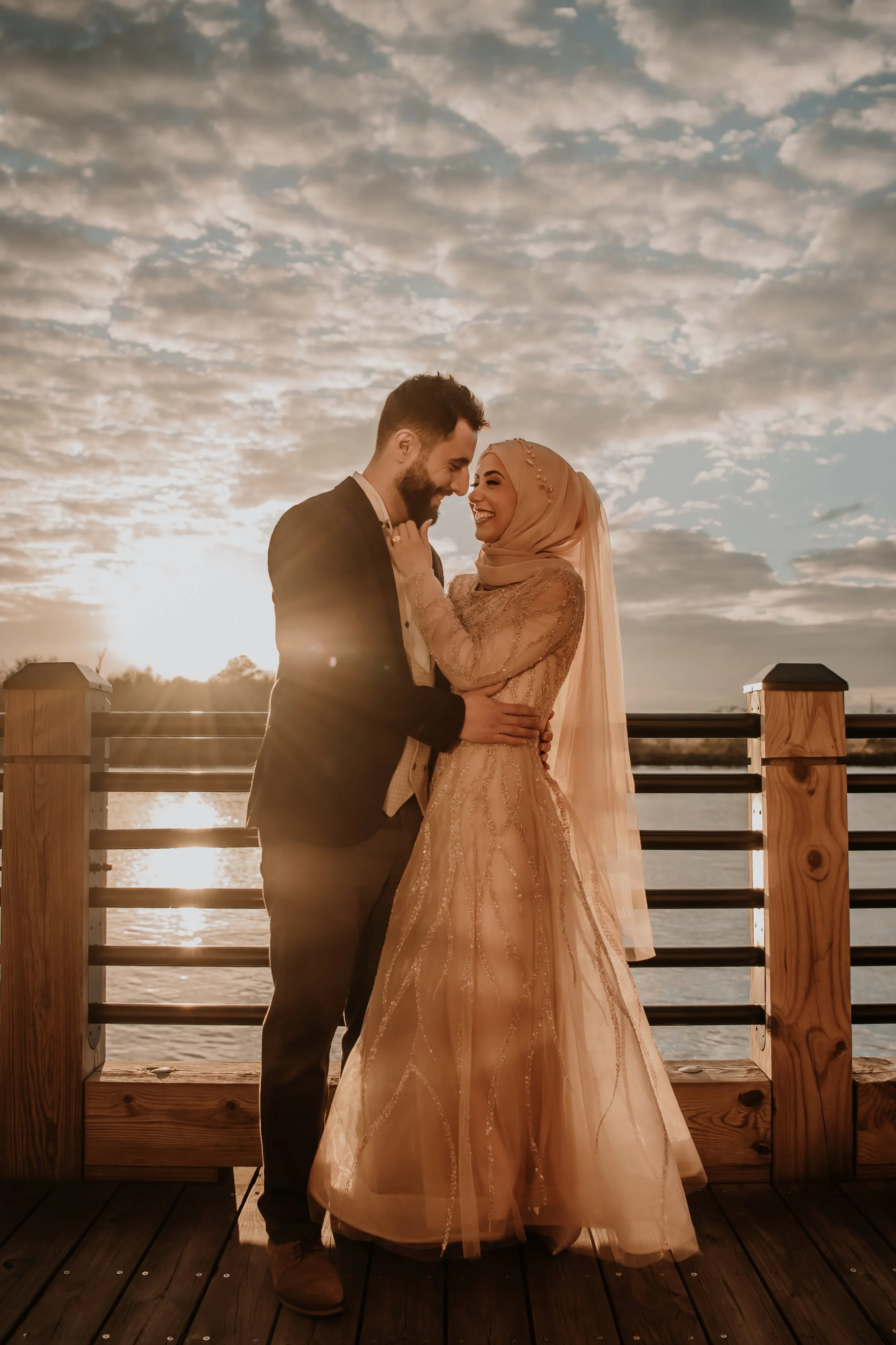 A bride and groom sharing a joyful moment on a wooden dock by the water at sunset.
