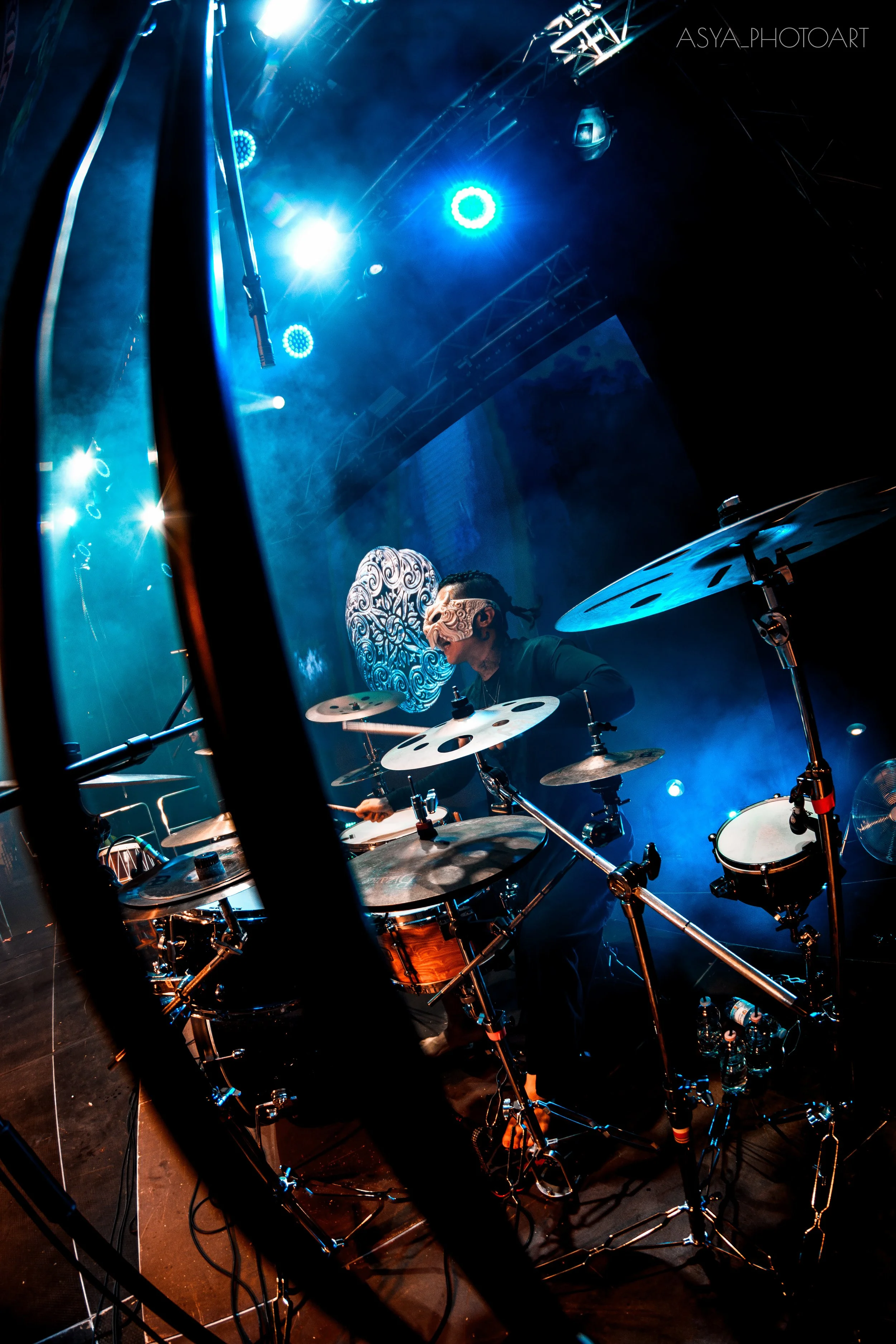 A drummer performing on stage with blue and white lights, wearing a face mask with intricate designs, surrounded by drums and cymbals, seen through a metal barrier.