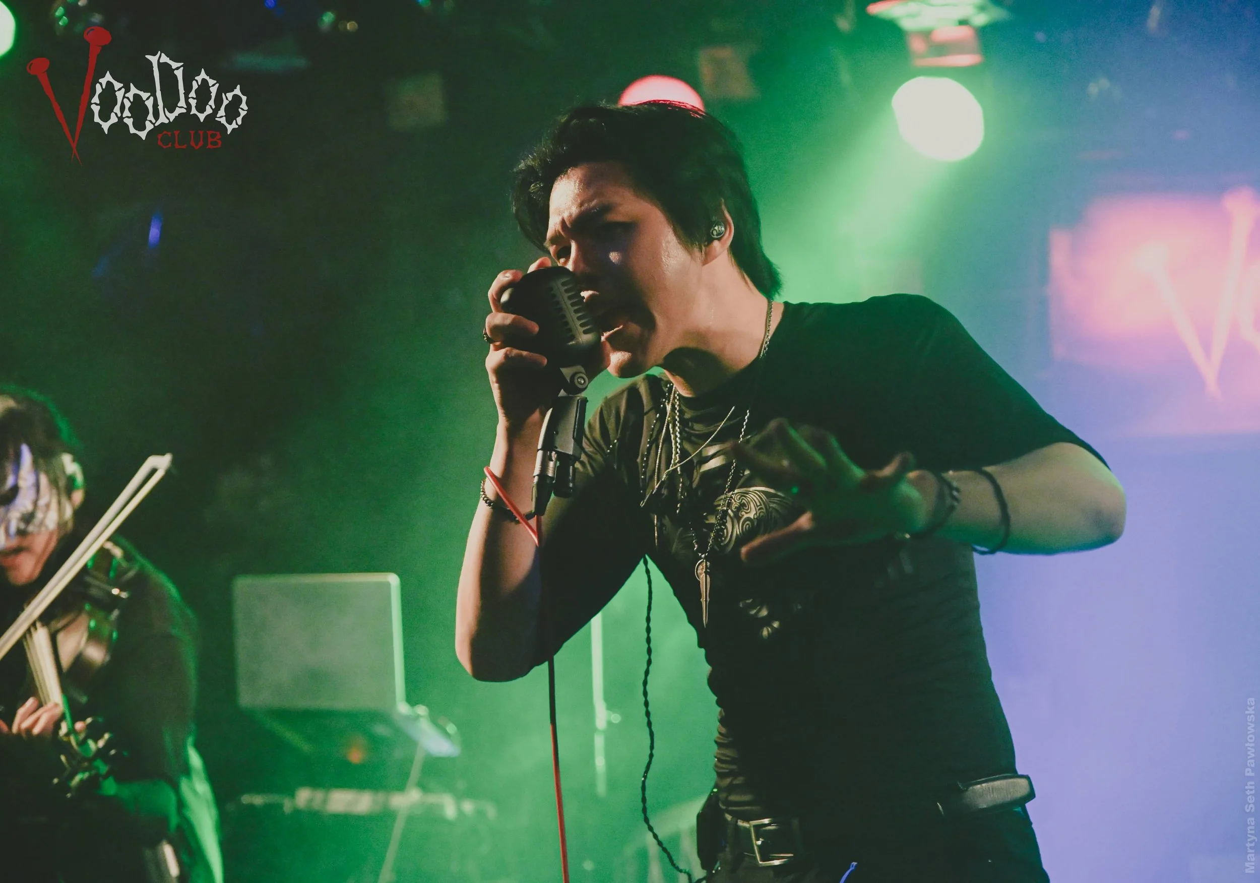A male singer performing with a microphone on stage surrounded by colorful stage lights in a club setting, with a logo reading 'Voodoo Club' in the top left corner.