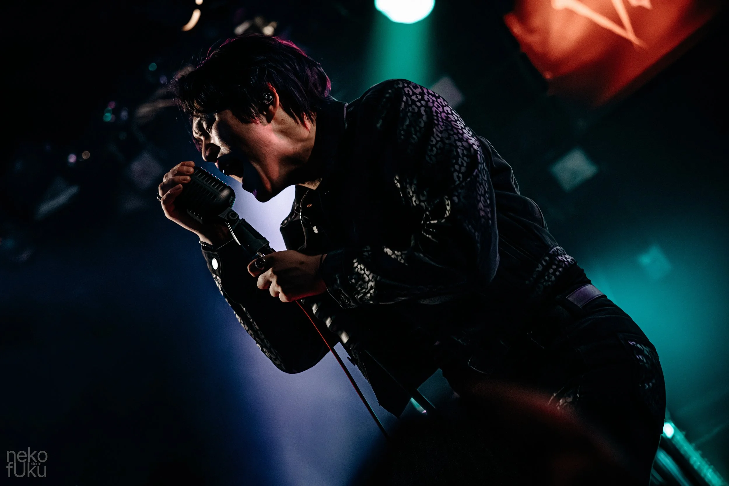 A singer passionately performing on stage, holding a microphone with a red cord, wearing a black and white patterned jacket, with colorful stage lighting in the background.