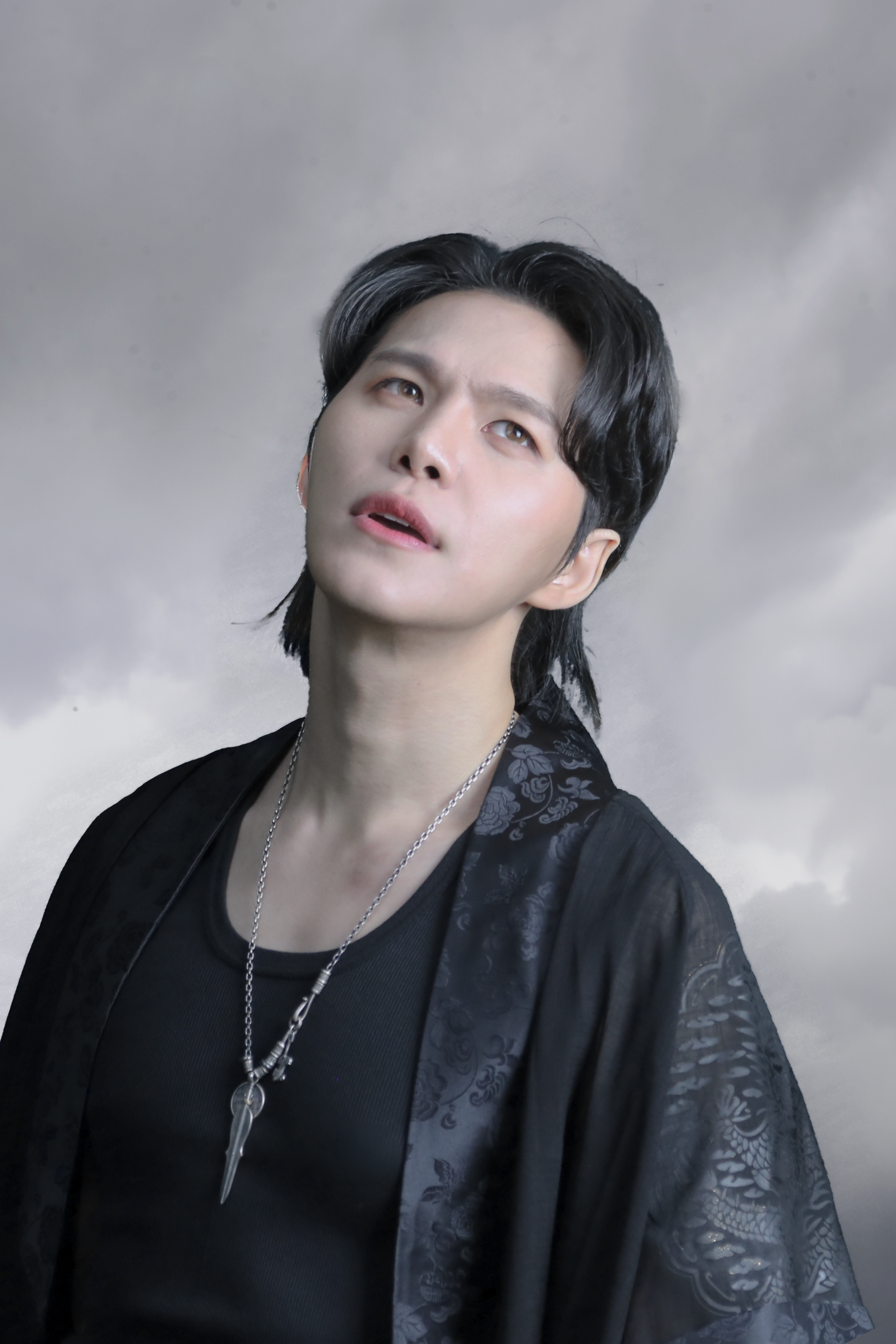 A young man with black, shoulder-length hair, wearing a black shirt and a black shawl with floral patterns, and layered necklaces, standing against a cloudy sky background.