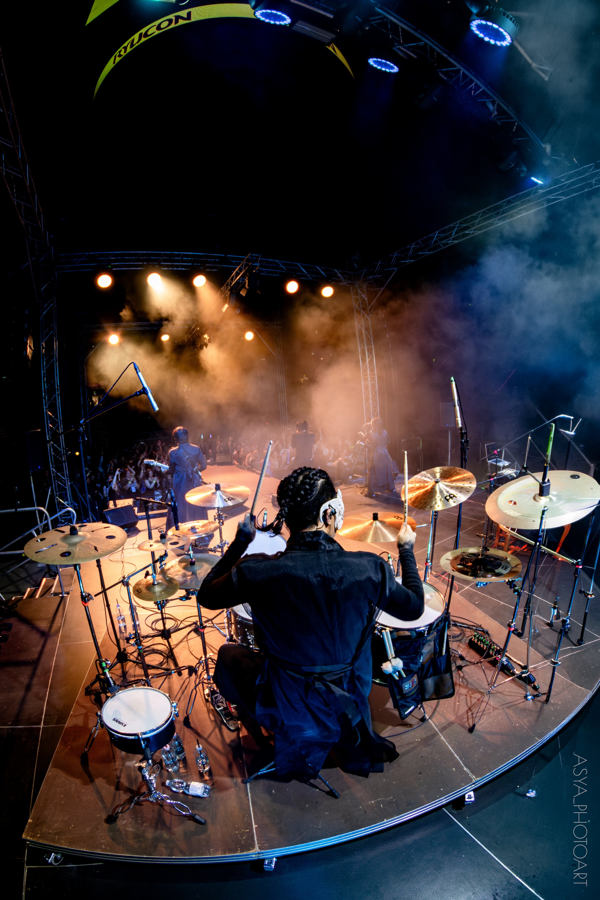 A drummer on stage during a live performance, seen from behind, with drums and cymbals in the foreground, stage lights overhead, and a crowd in the background.