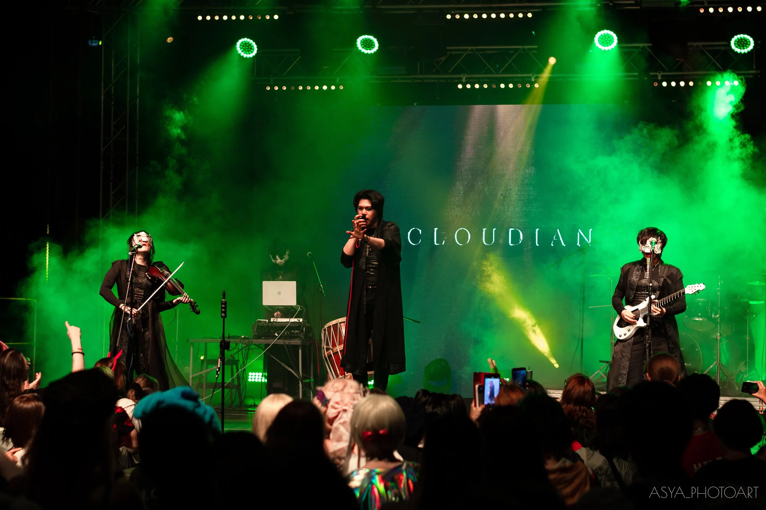 Musicians performing on a stage with green lighting, including a violinist, a singer, and a guitarist, in front of an audience.
