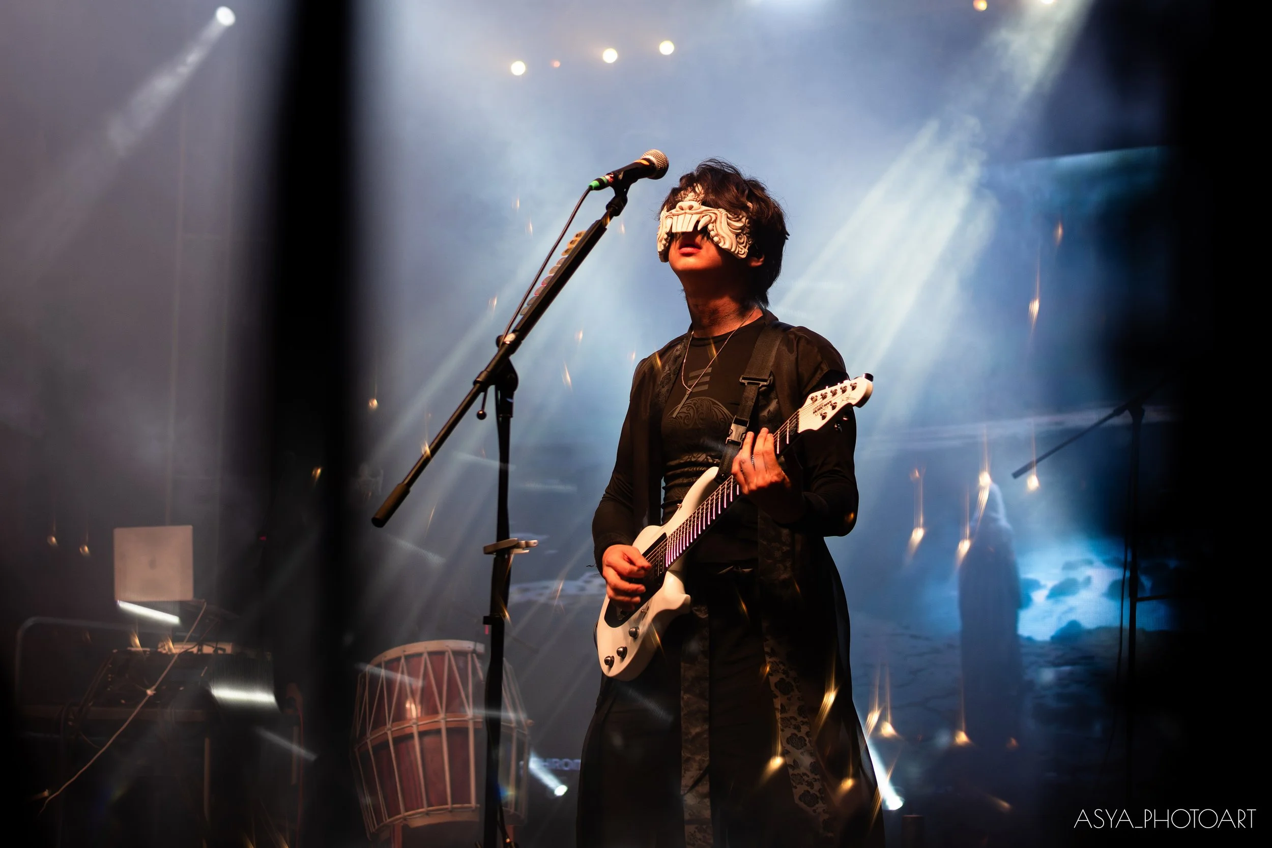 A musician playing guitar on stage, wearing a decorative blindfold mask and dark clothing, with stage lights and smoke effects in the background.