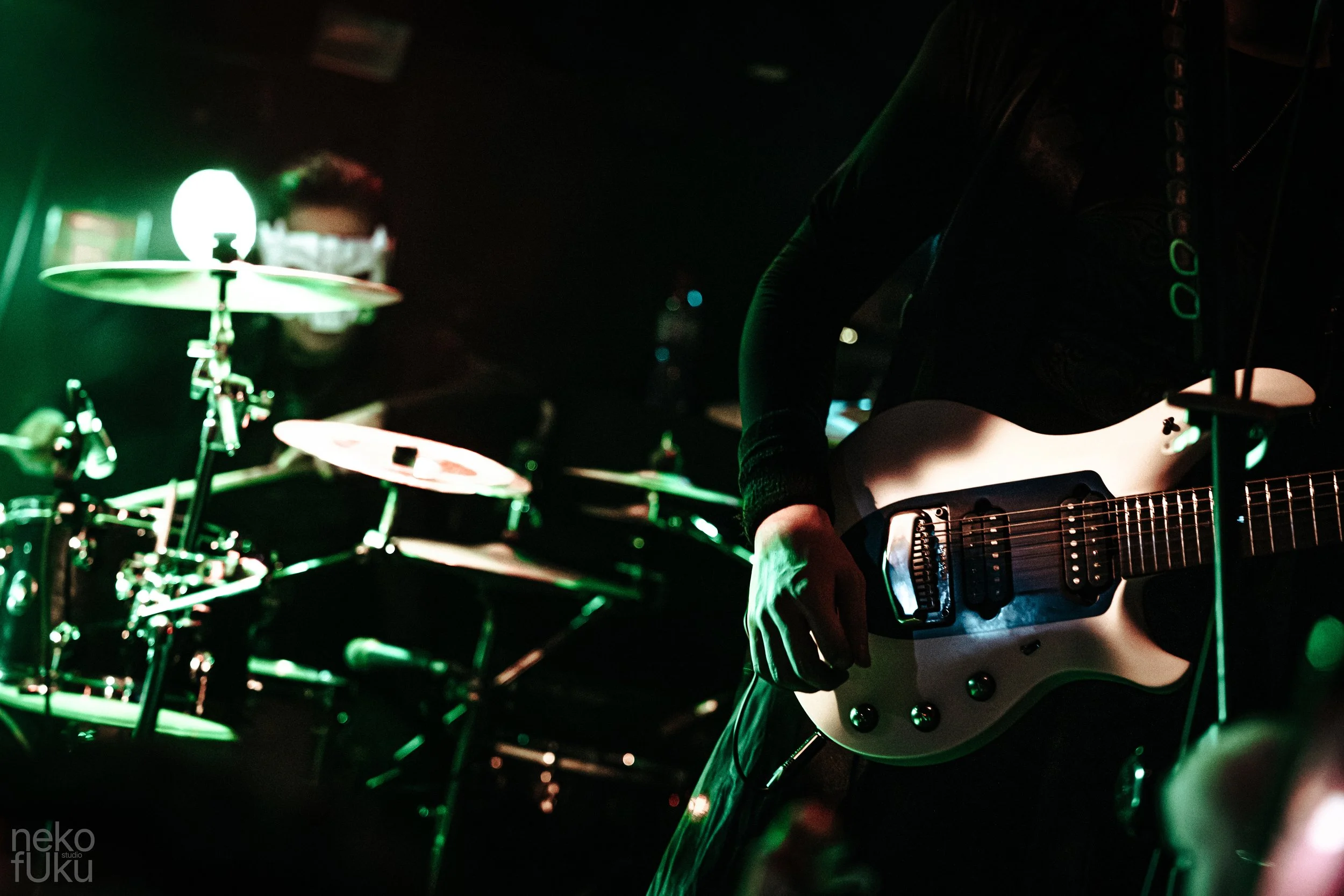 A musician playing an electric guitar in a dimly lit setting, with a drummer in the background.