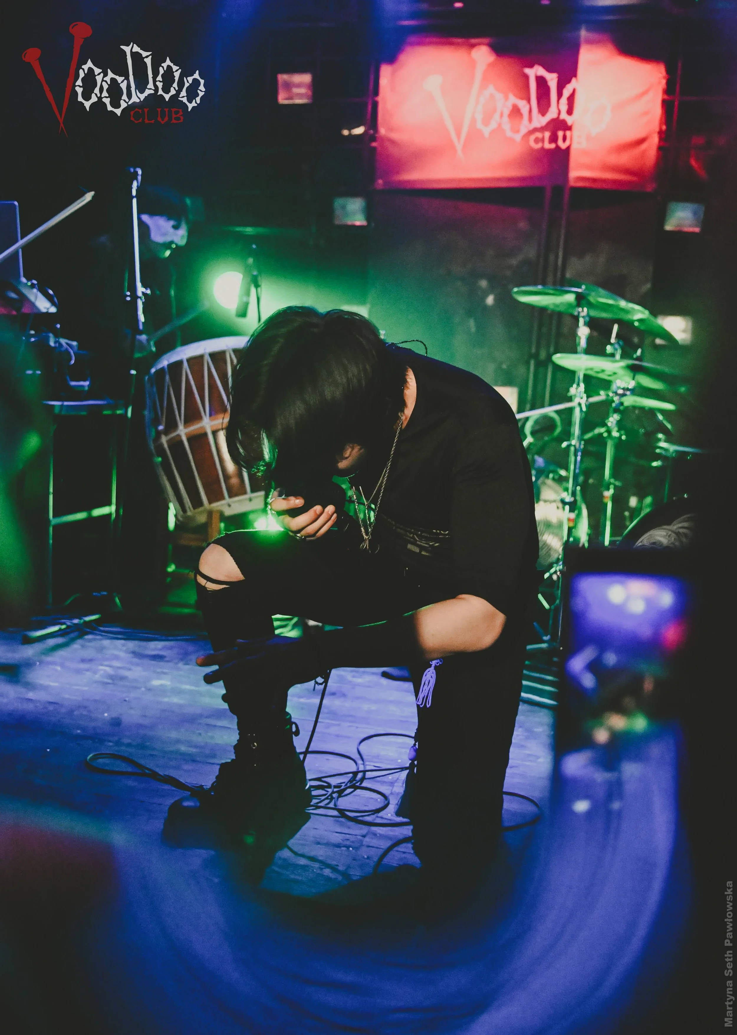 Person kneeling on stage with their head down, holding a microphone, with musical instruments and a 'Voodoo Club' sign on the wall behind them, illuminated by colorful stage lights.