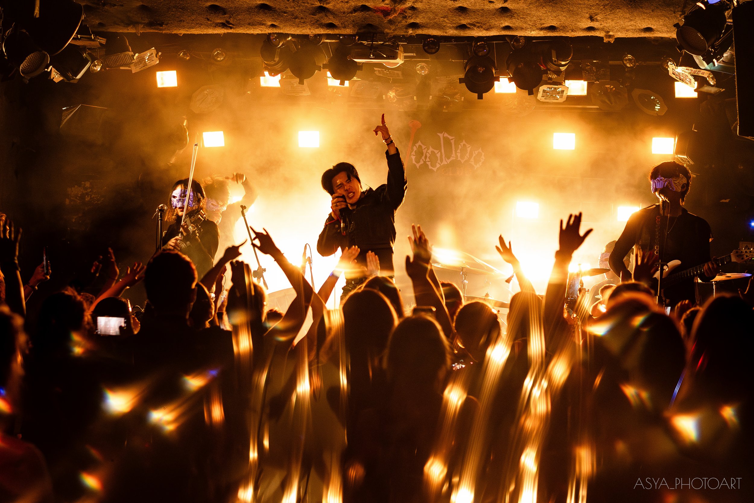 A lively concert scene with a band performing on stage under warm, bright lights, and an energetic audience with raised hands in front.
