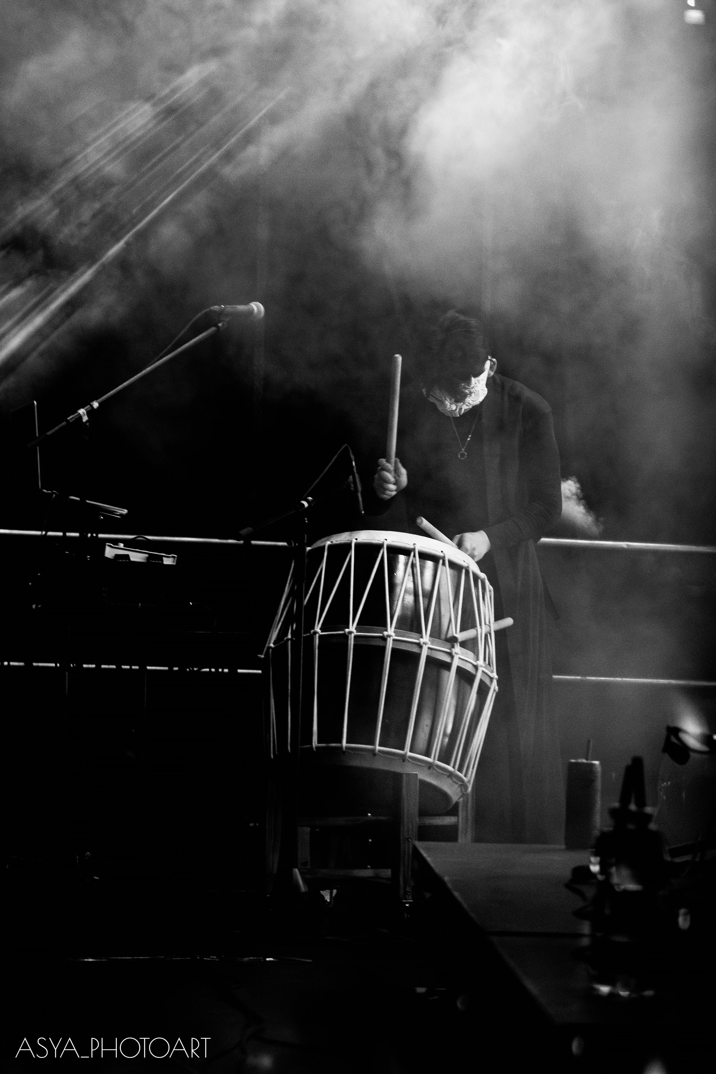 A person with curly hair and a face mask playing a traditional drum with a stick in a dark, smoky environment, with stage lights creating a misty atmosphere.