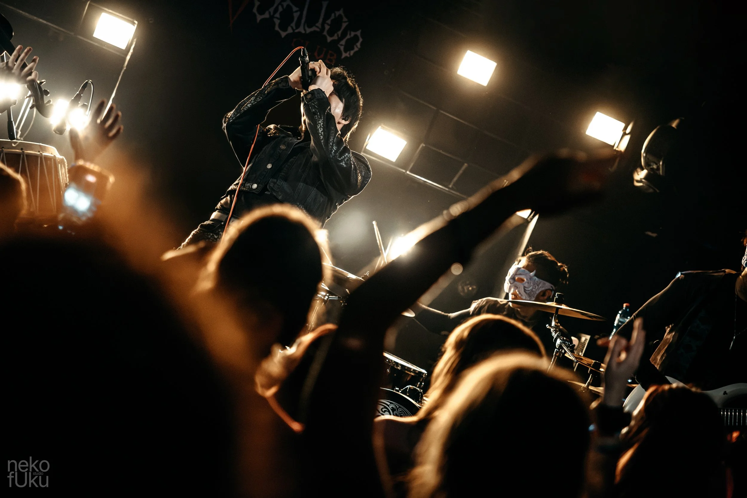 A performer singing on stage with hands raised above head, facing a crowd at a concert with bright stage lights and a dark background.