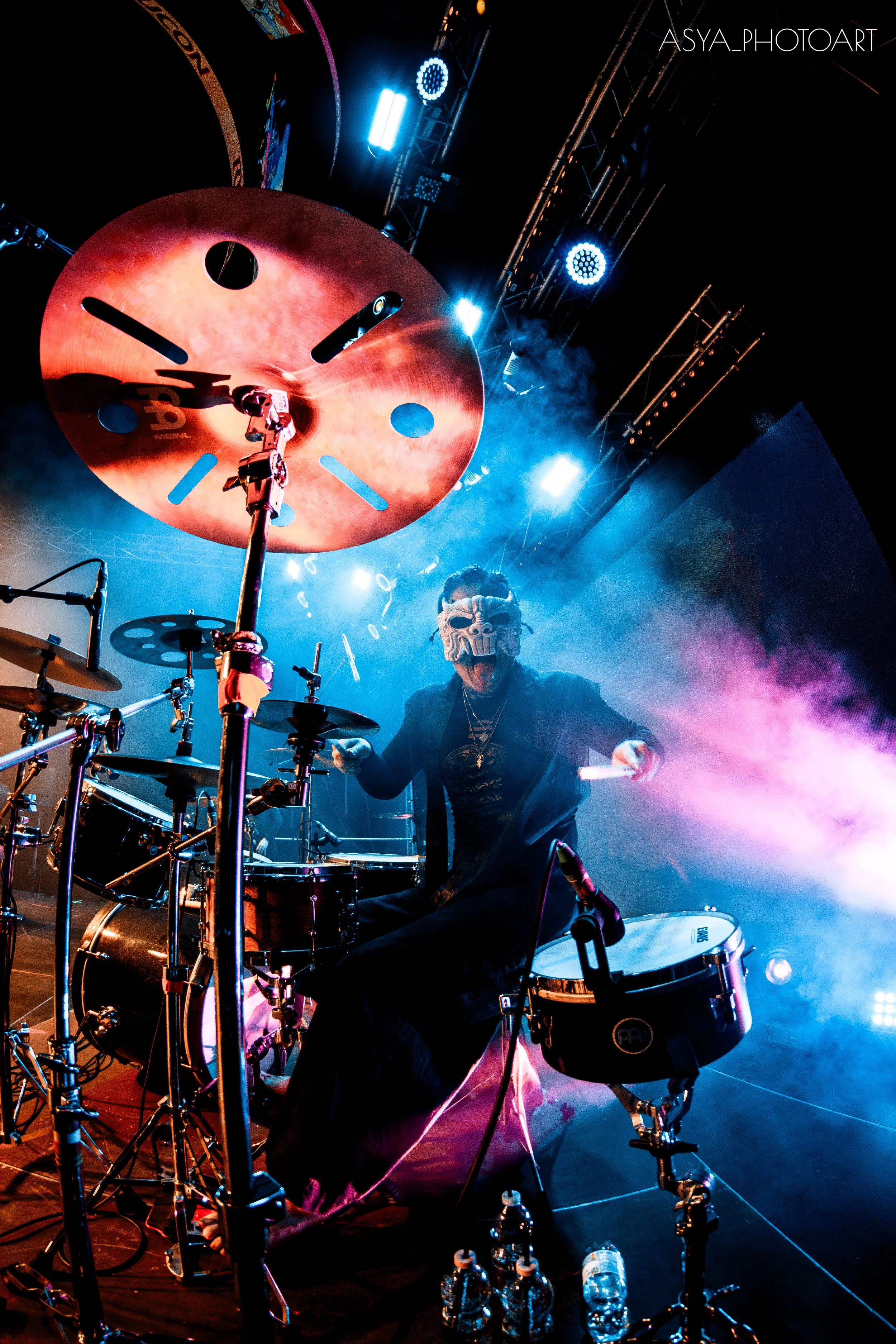 A musician wearing a tribal mask playing drums on stage with colorful lights and fog.