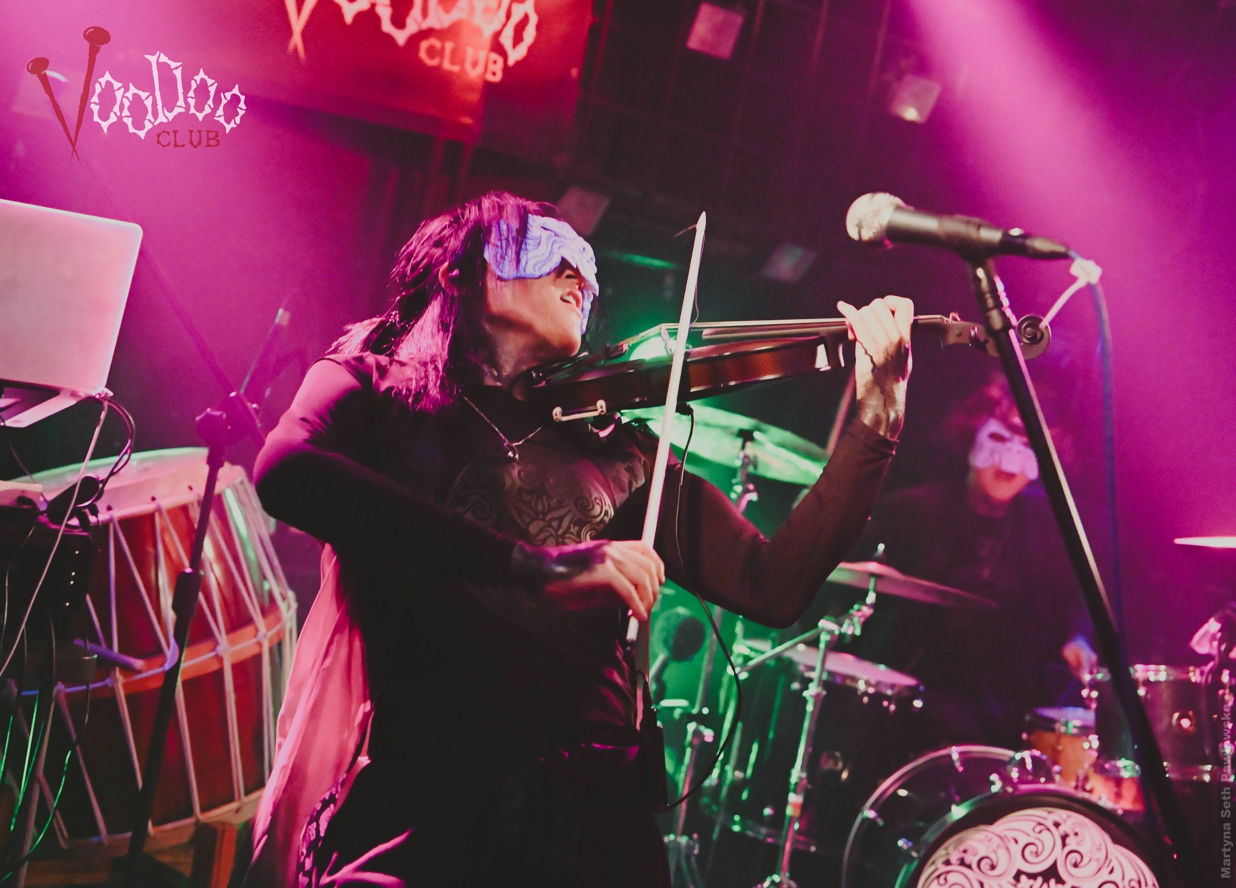 A woman wearing a blindfold playing a violin on stage at a club, with a microphone nearby and colorful stage lighting.