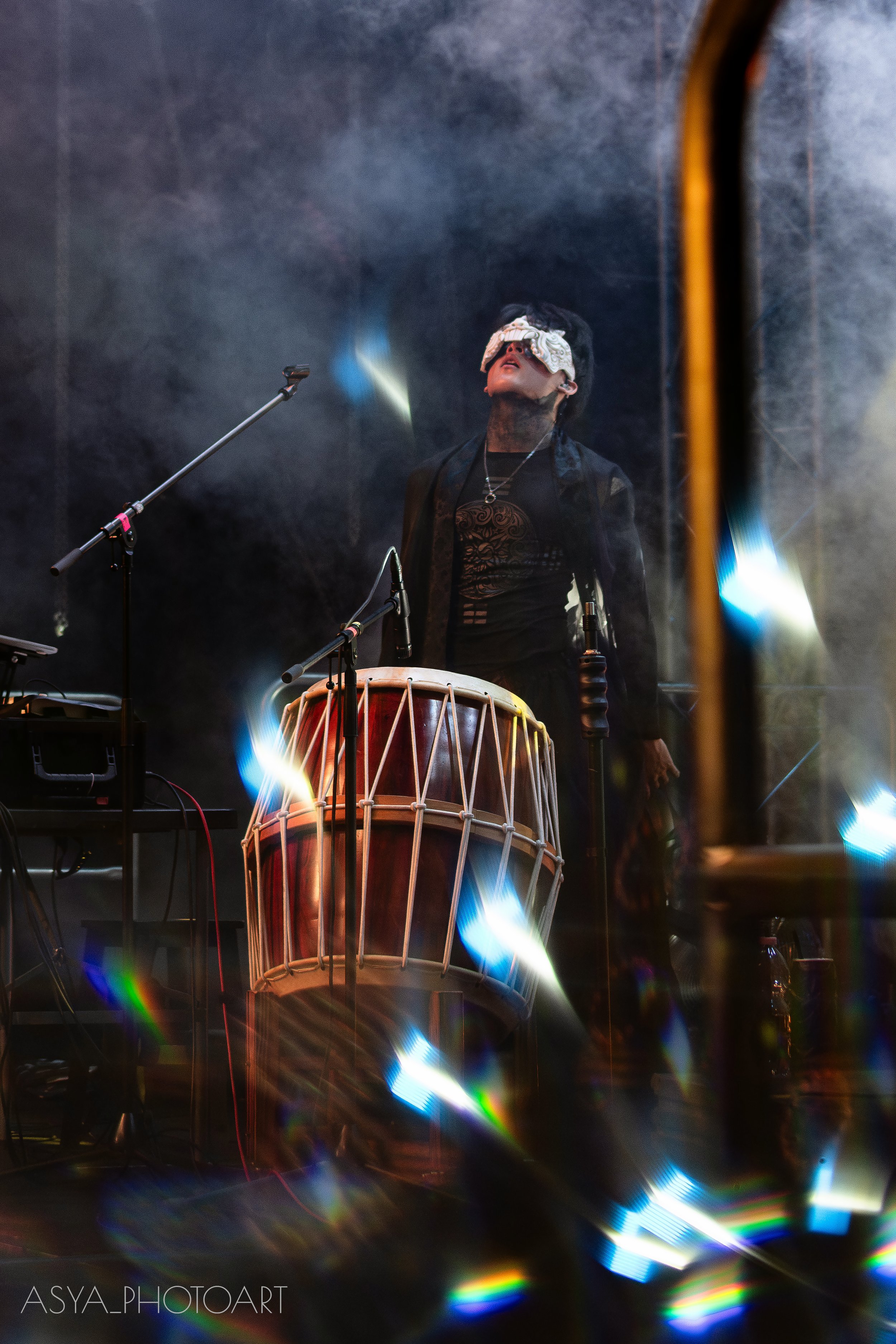 Person wearing a blindfold, standing behind a large drum with colorful strings, positioned in front of a dark, smoky background with stage lights.