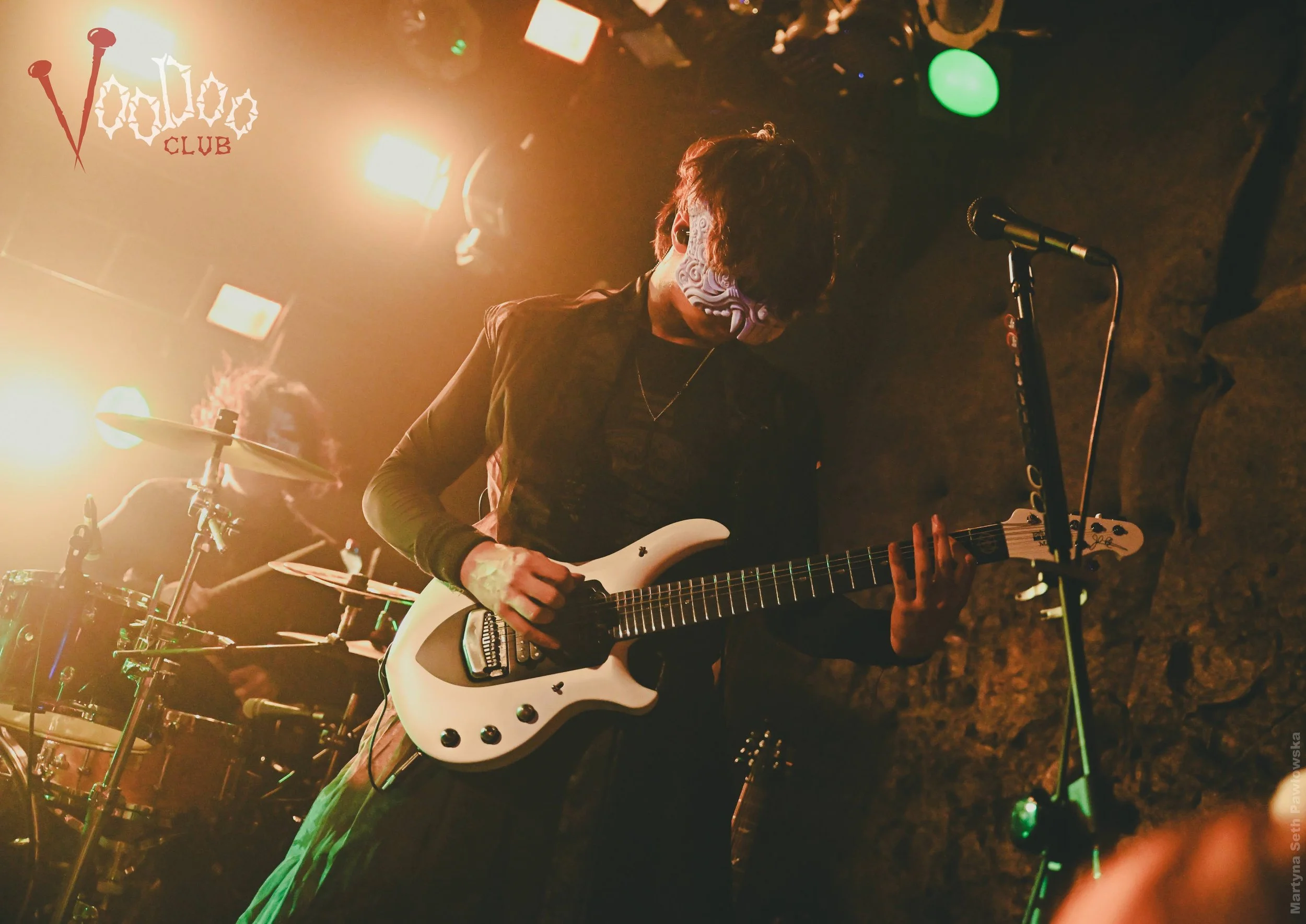 Musician playing an electric guitar on stage with a microphone in front. Stage lights and a drum set are visible in the background at a rock concert or nightclub.