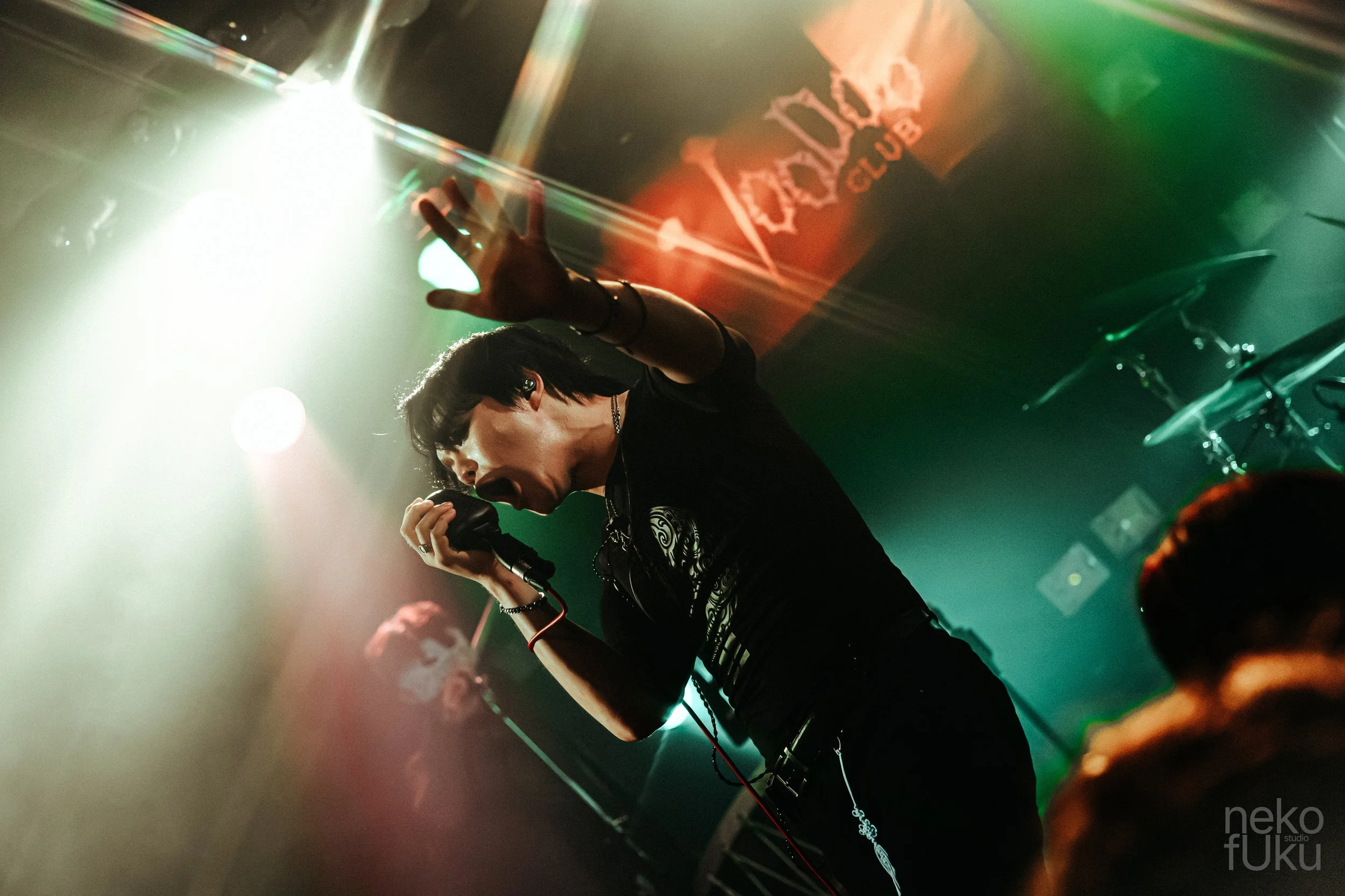 A singer performing on a dimly lit stage, holding a microphone with left hand, wearing black clothing, with colorful stage lights and a banner in the background.