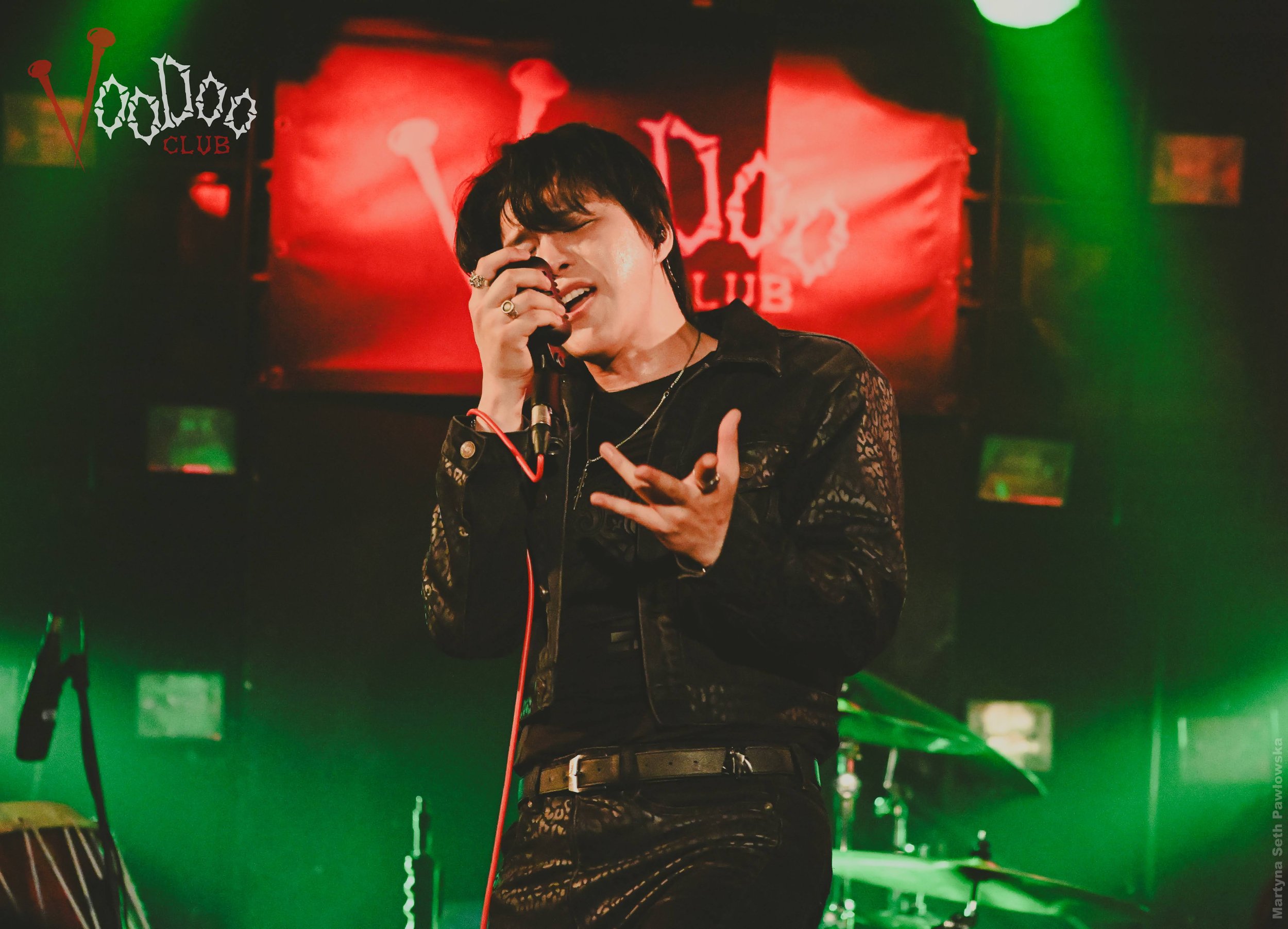 A singer with black hair performing on stage at a nightclub, with a red and green lit background featuring a "Voodoo Club" sign.