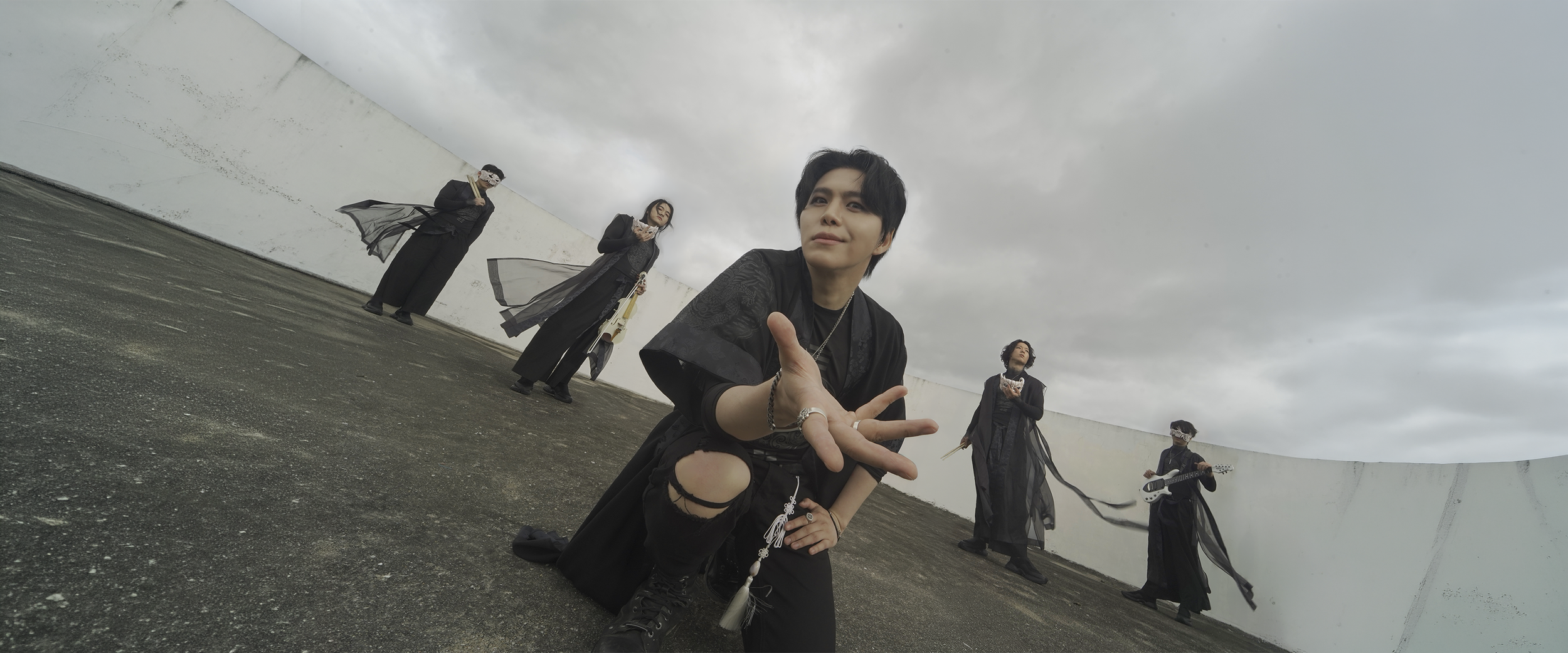 Group of five people on a rooftop, dressed in black and gothic clothing, with one person kneeling in front and making a peace sign, others standing with musical instruments and drinks, under a cloudy sky.