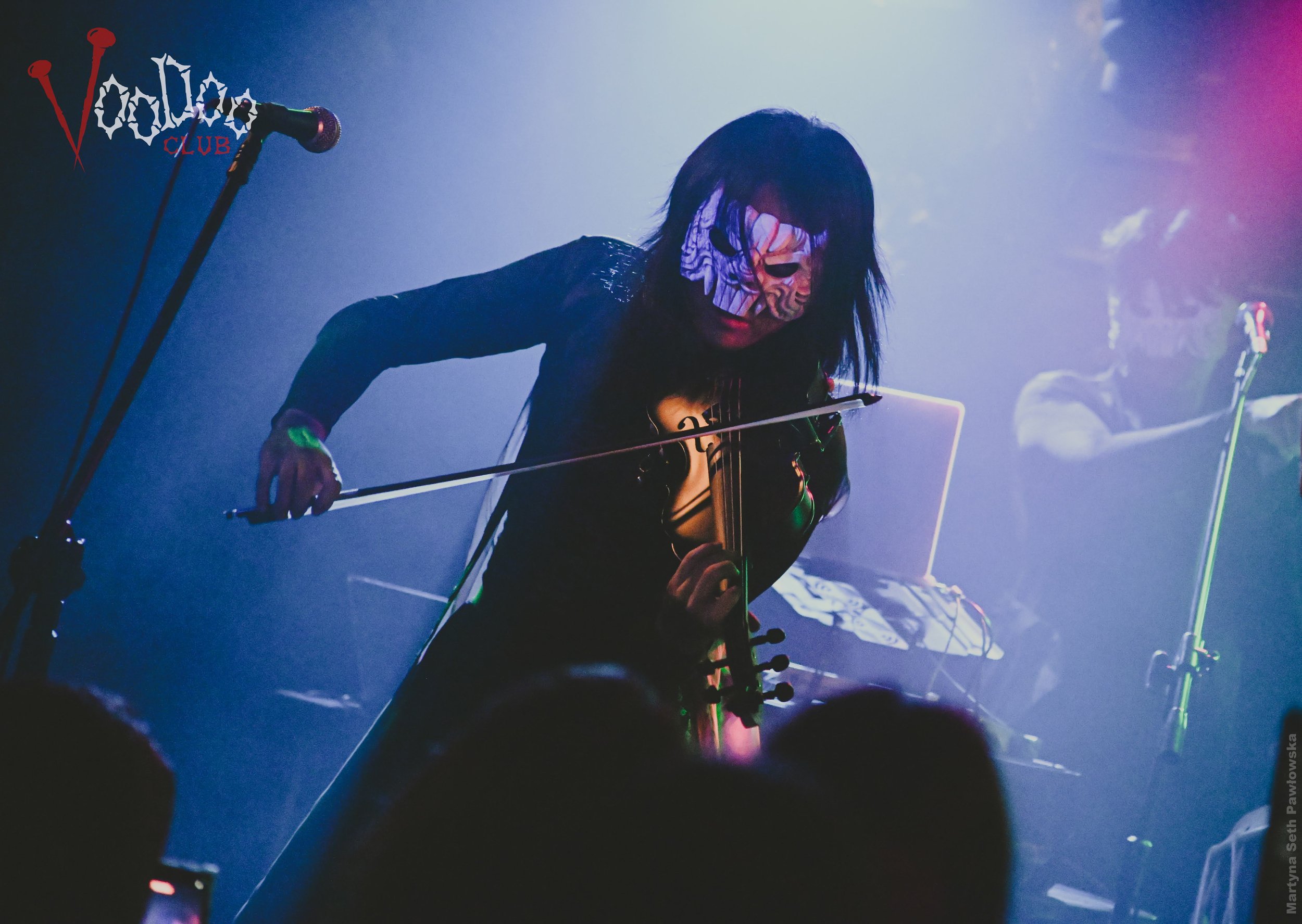 Person with long dark hair playing a black electric violin on stage, wearing a face mask with a colorful design, illuminated by stage lights, in front of a keyboard and equipment, with a microphone stand nearby. The background shows a logo for Voodoo