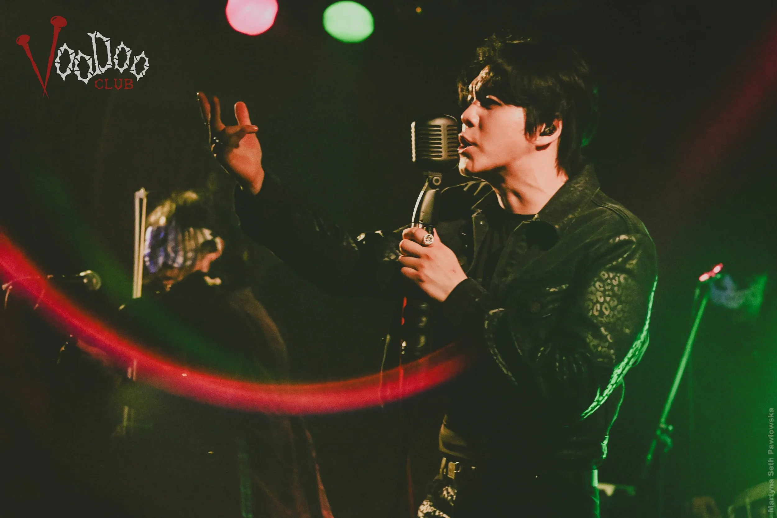 A male singer with dark hair wearing a leather jacket, performing with a microphone on stage at a club called Voodoo Club, with colored lights in the background.