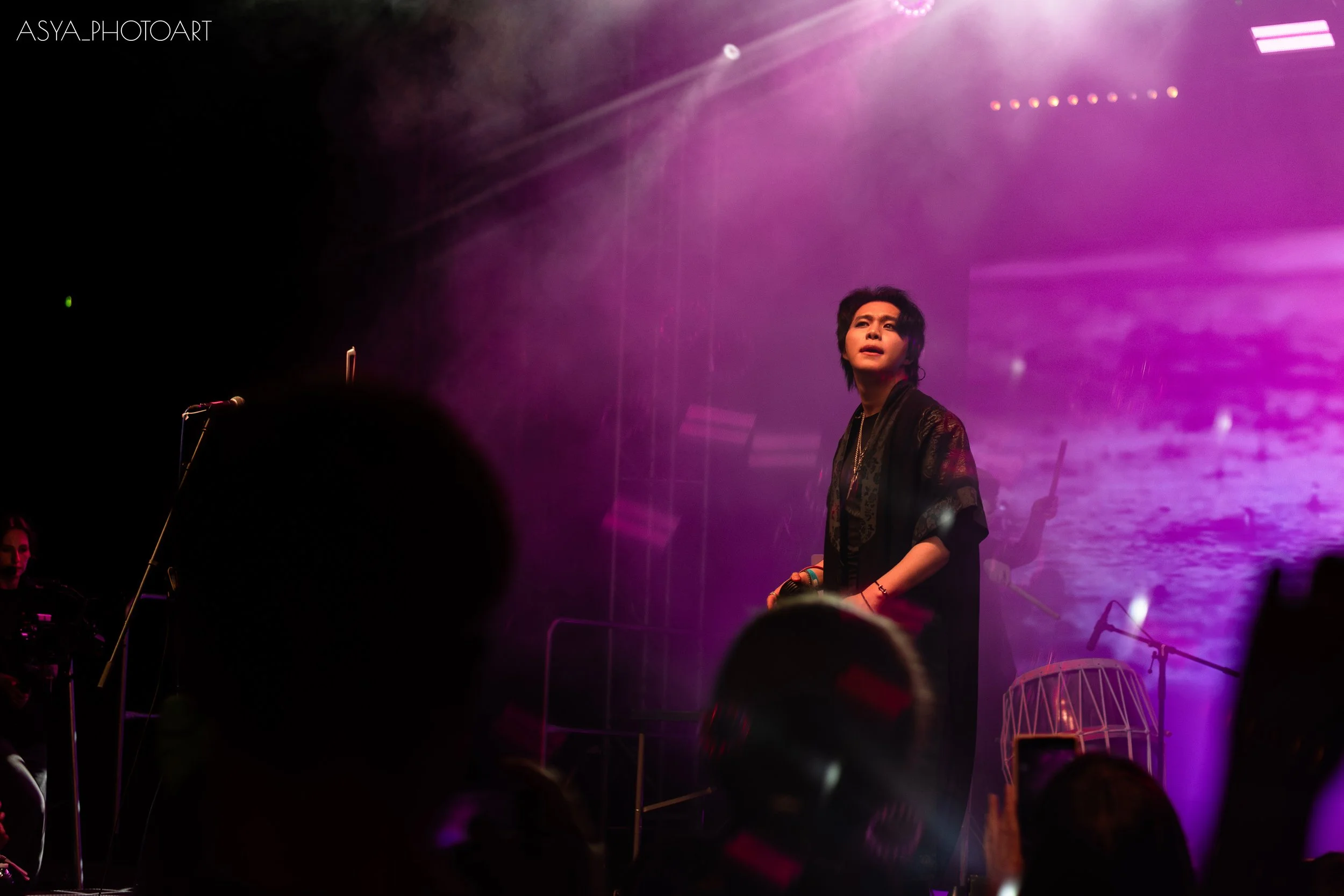 A person with dark, shoulder-length hair stands on stage illuminated by purple and pink stage lights. There are musical instruments and microphones visible, suggesting a live performance or concert setting. Audience members are faintly visible in the