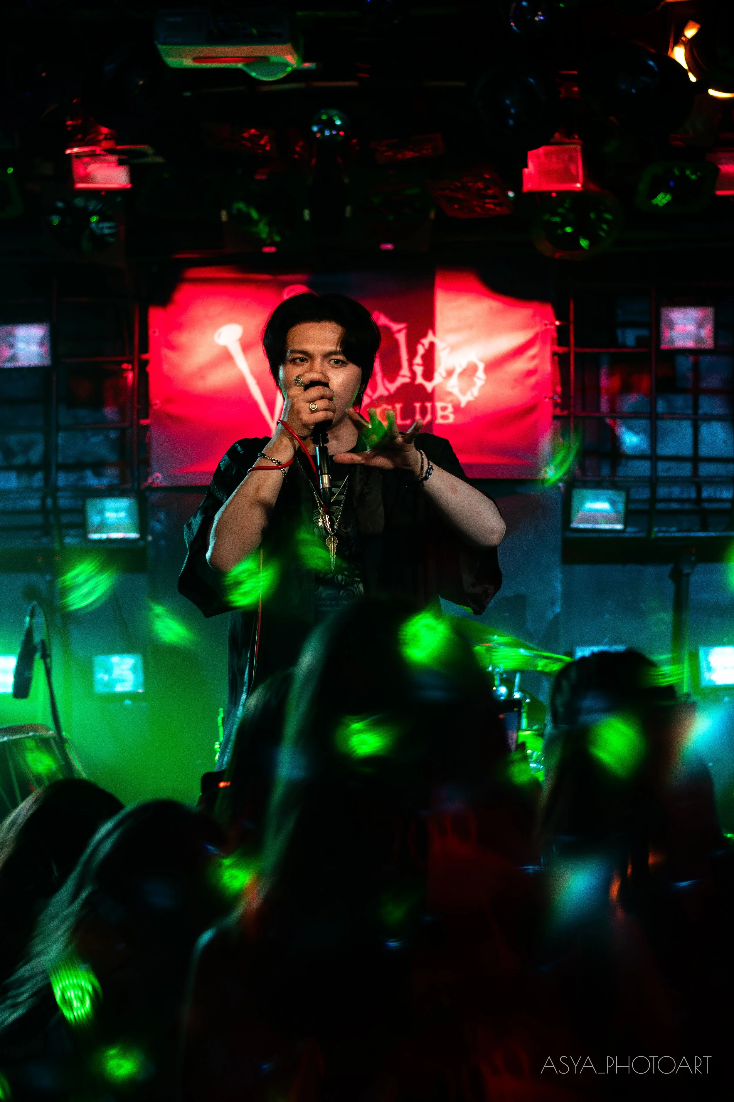 A male singer performing on stage with a microphone, in a club with colorful lighting. The background displays a sign that says 'CLUB'.