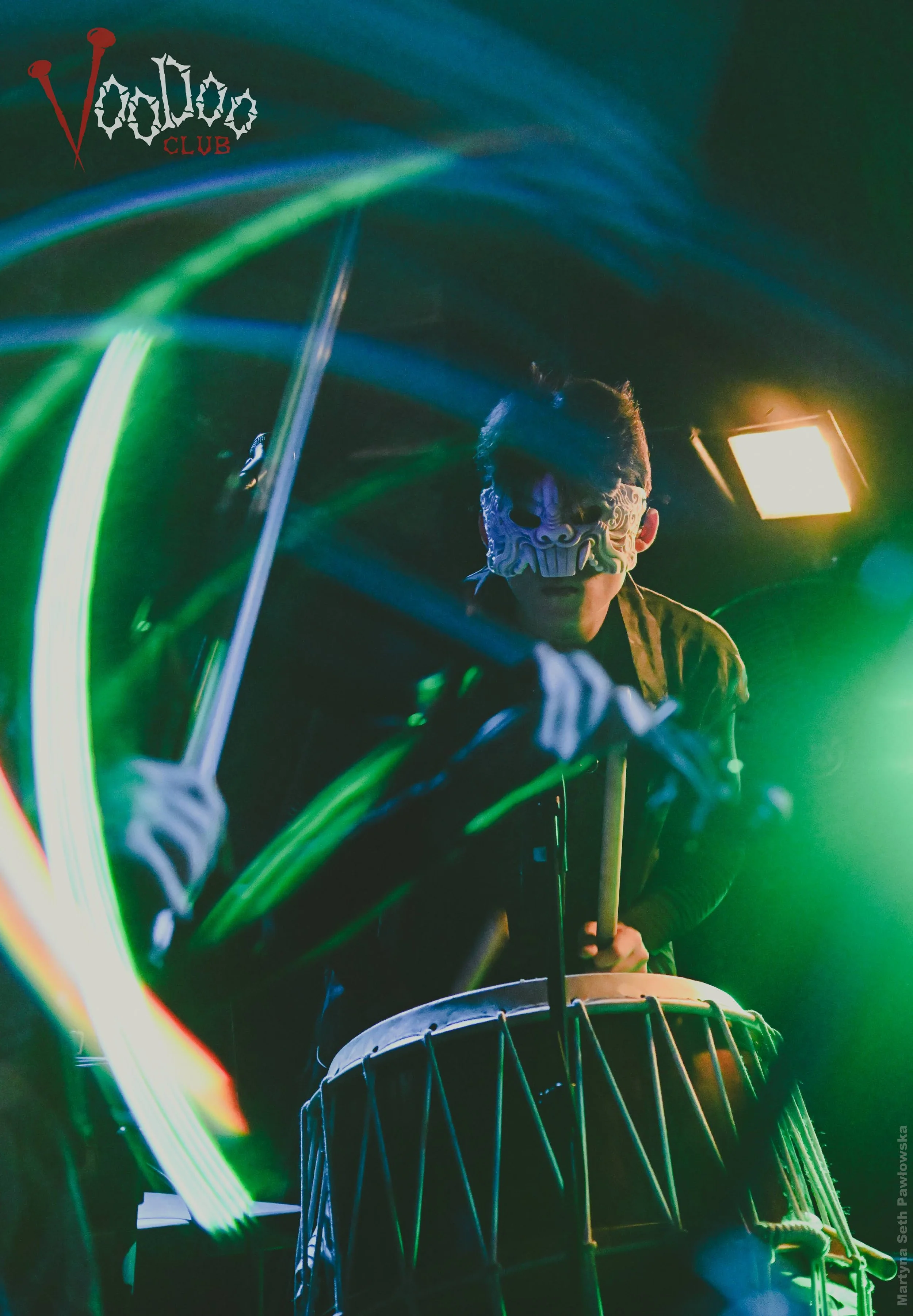 Person wearing a decorative mask playing a drum in a dimly lit setting with colorful light streaks.