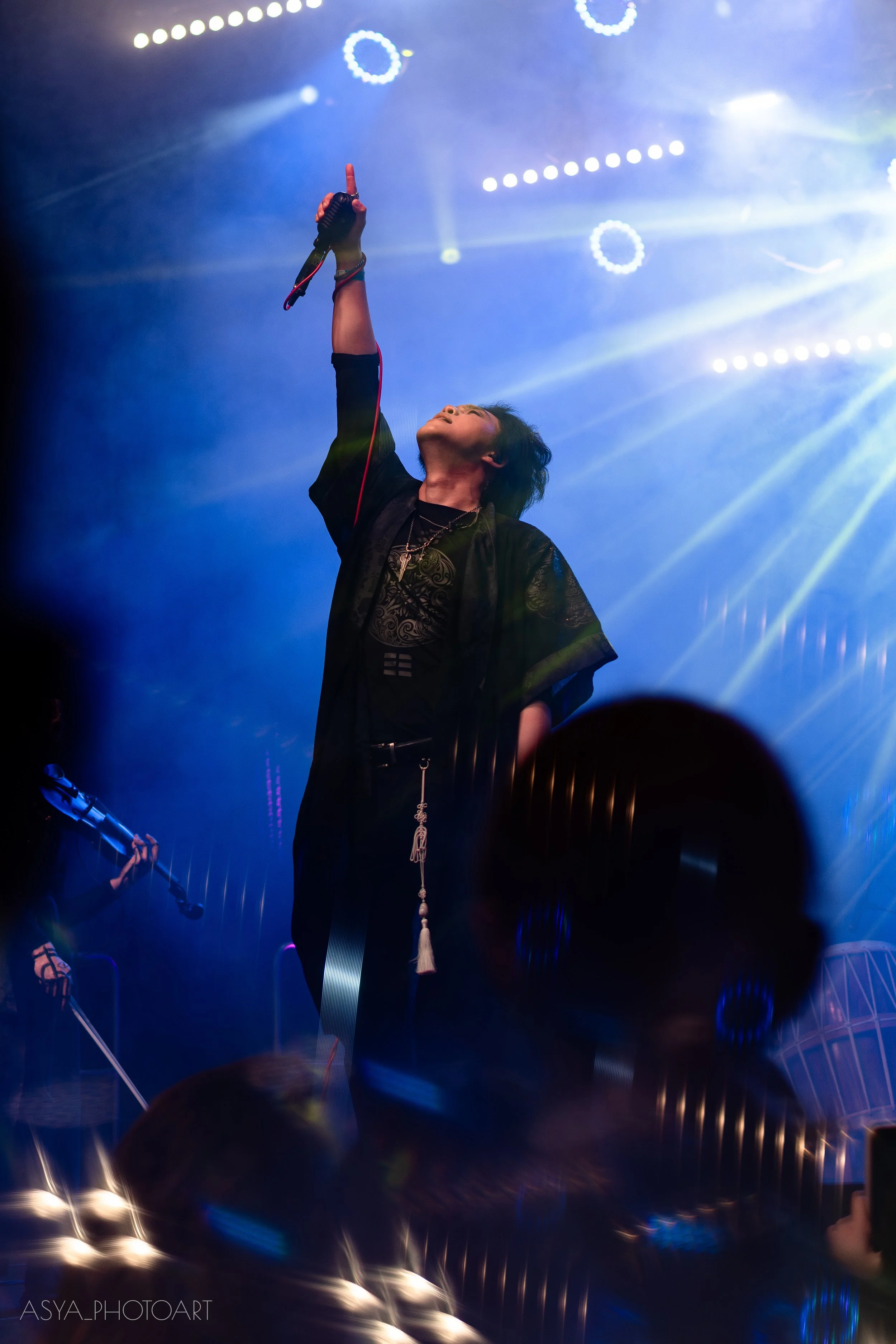 A person on stage holding a microphone high in the air, illuminated by colorful stage lights and beams, with a dark background and silhouettes of others playing instruments.